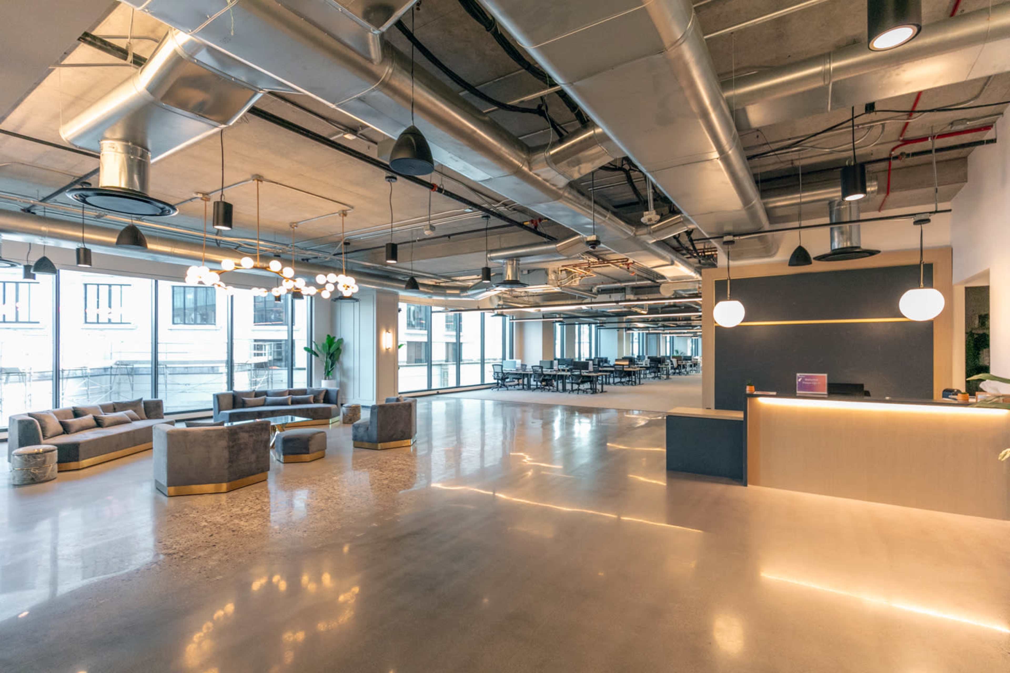 A modern open office space with large windows, polished concrete floors, and various seating areas, alongside a reception desk.