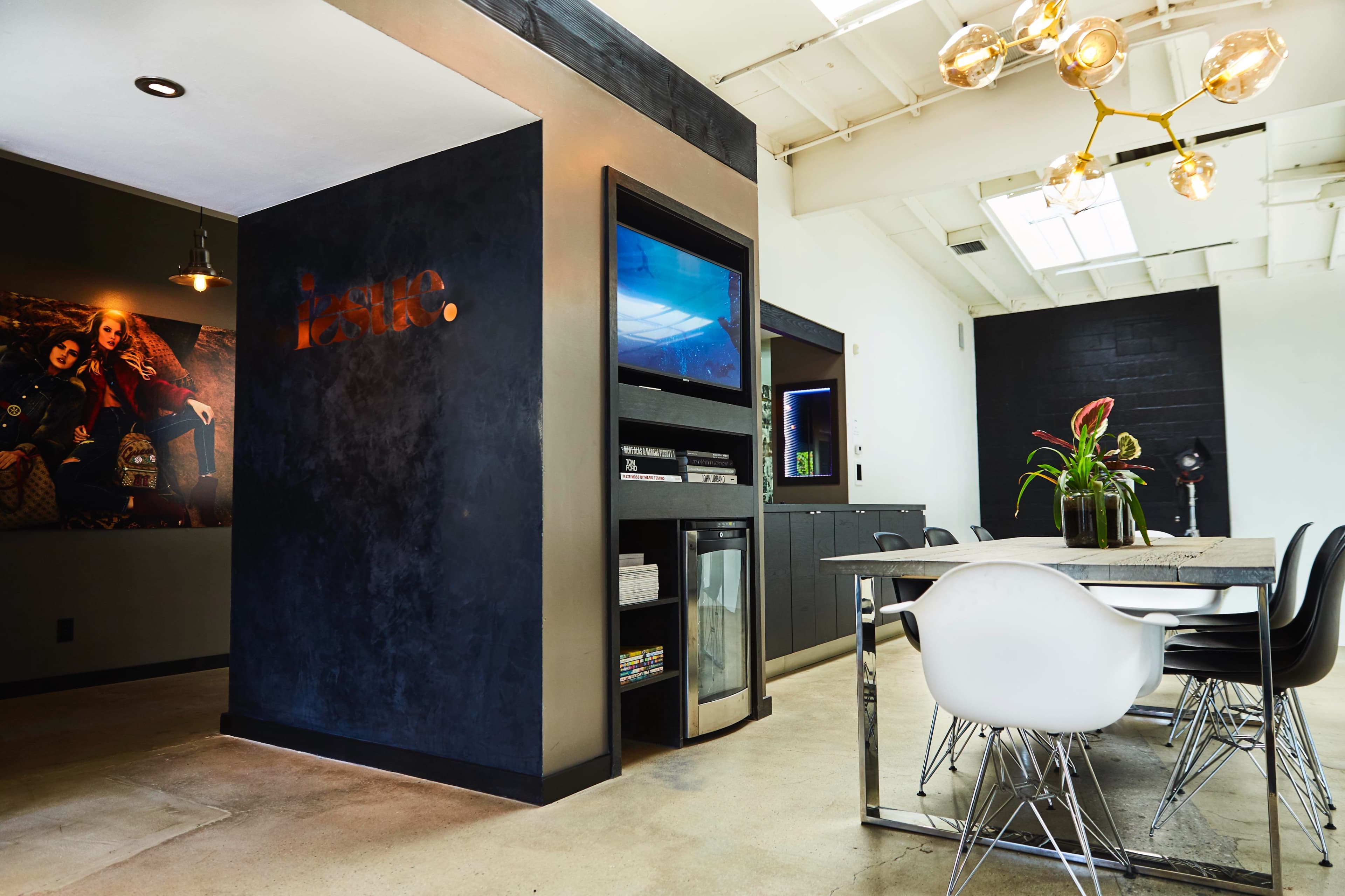 A modern office space features a dining table, a television display, and large windows with plants arranged on a central table.