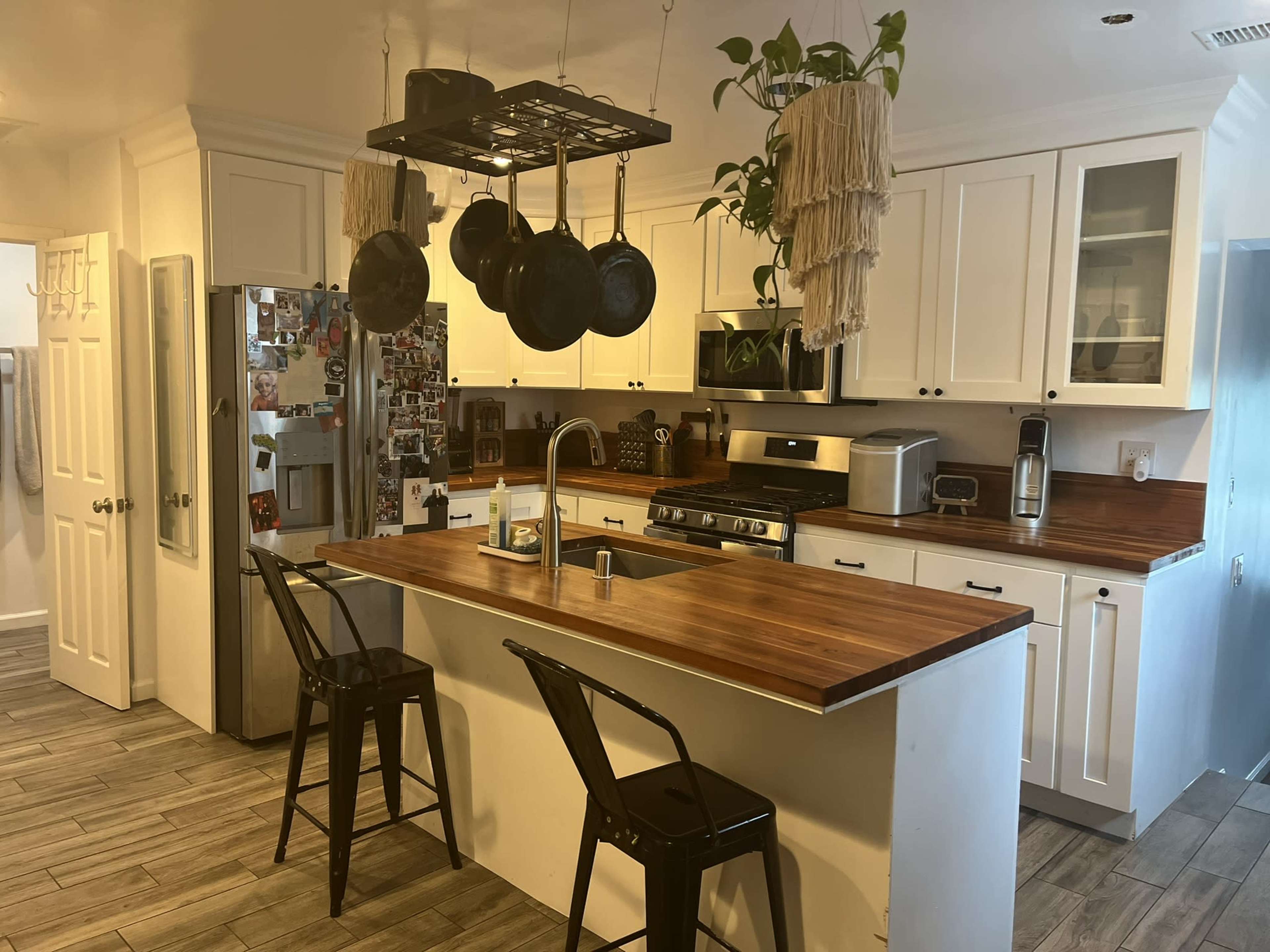 A modern kitchen features white cabinetry, a wooden countertop island with two black stools, and a pot rack hanging above.