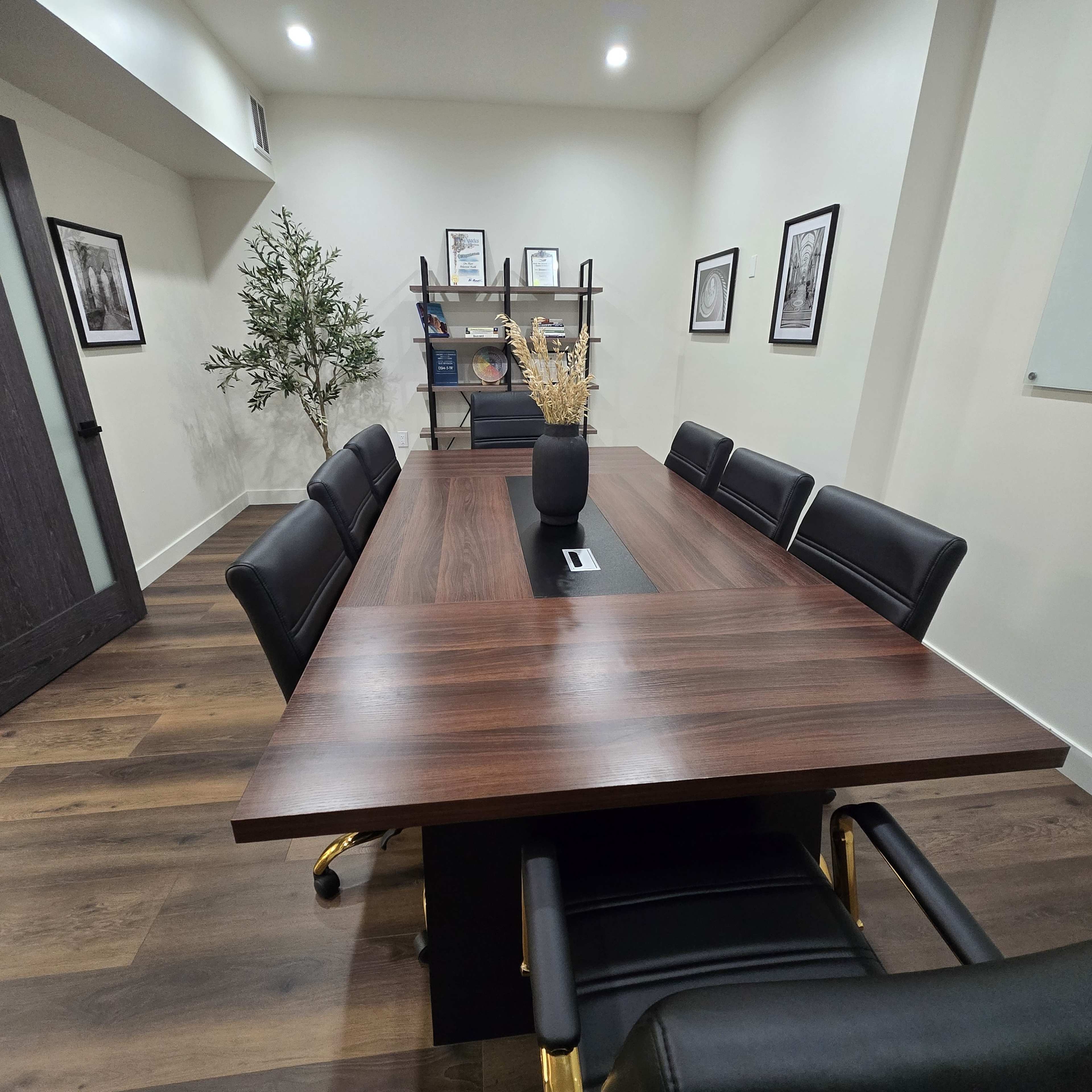 The image shows a conference room with a large wooden table, black leather chairs, and a bookshelf containing framed pictures and decorative items.