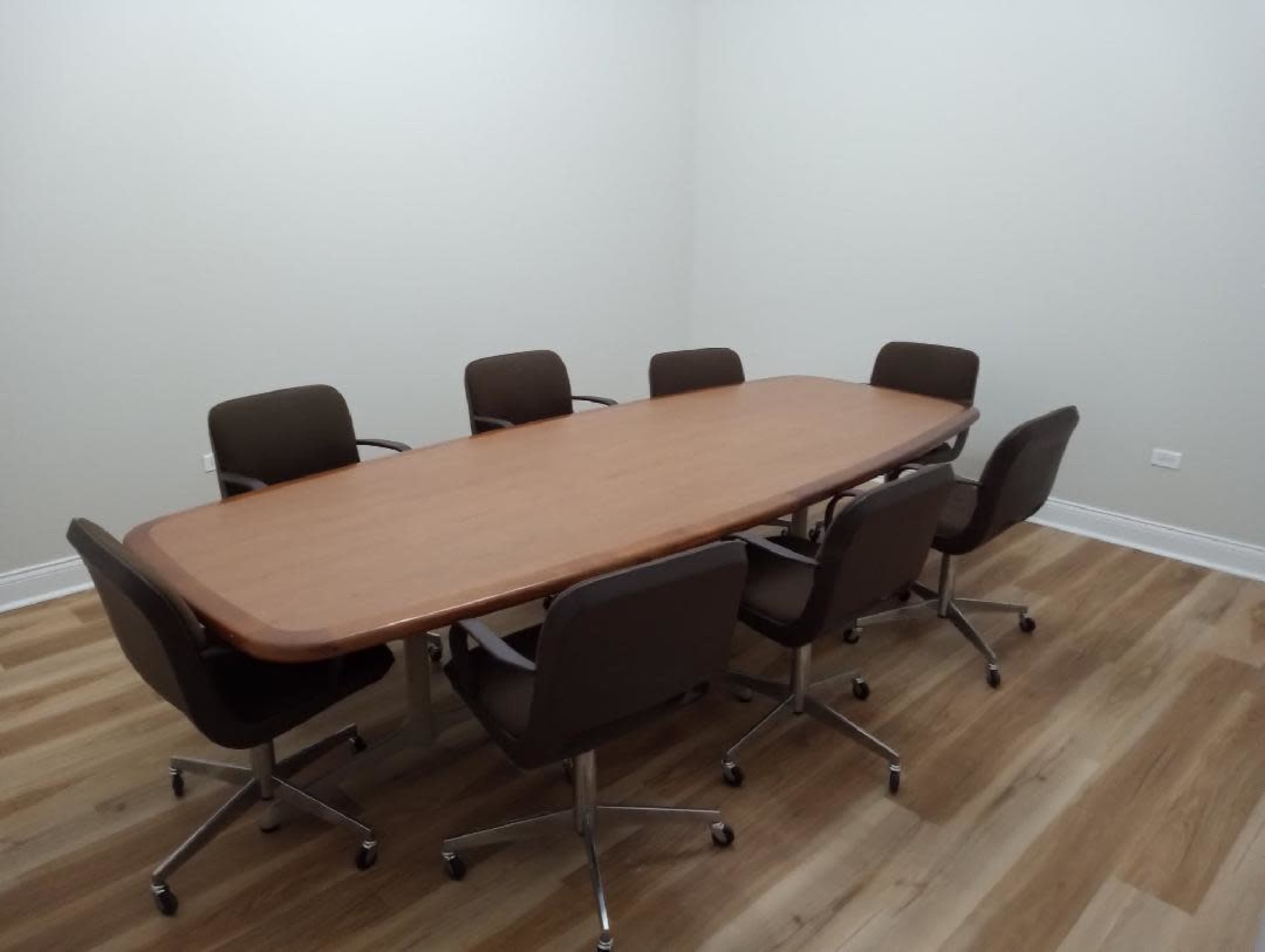A large wooden conference table surrounded by six rolling chairs is positioned in a sparsely decorated meeting room with a light-colored wall and wooden floor.