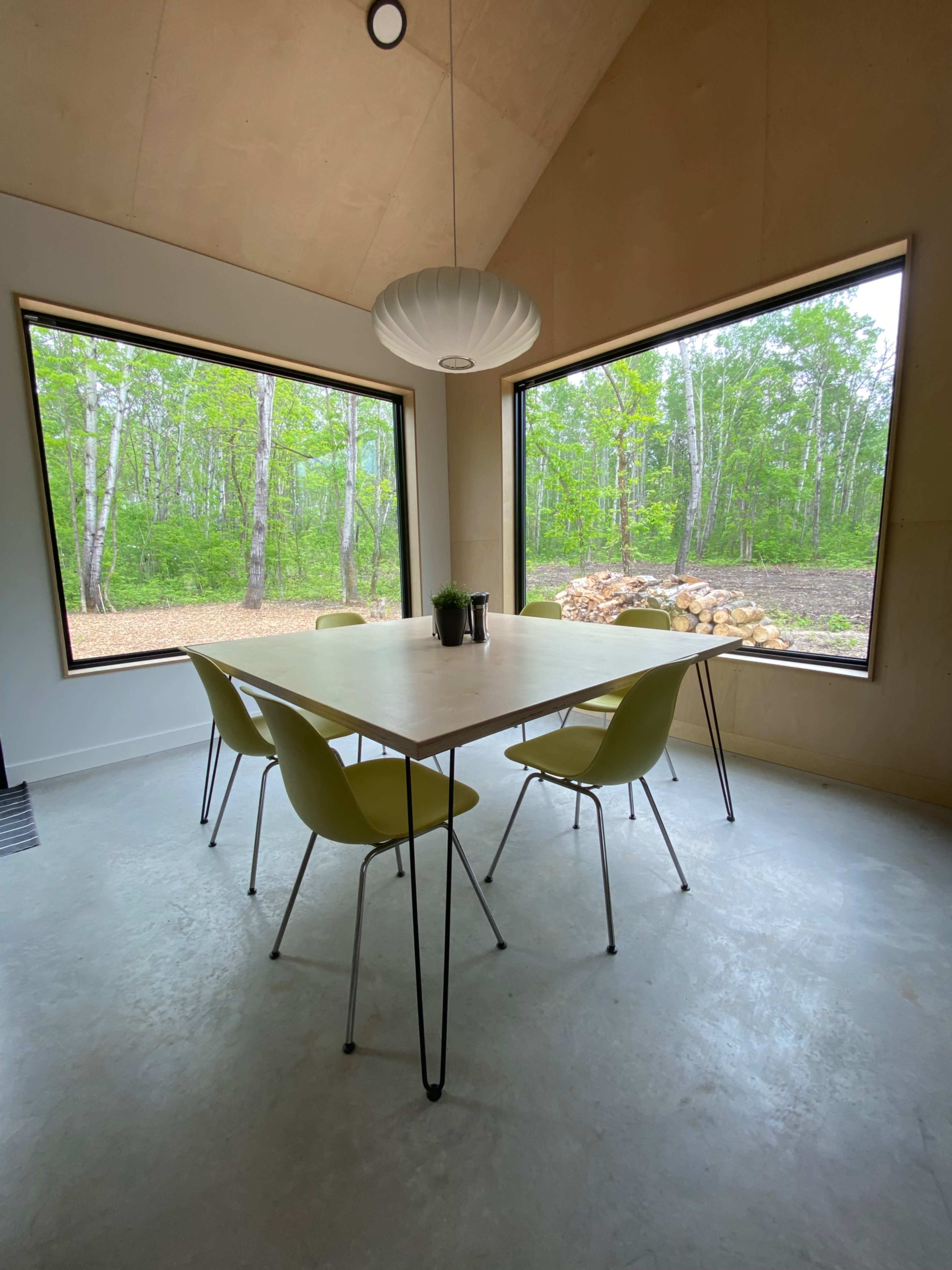 A minimalist dining area features a light wood table surrounded by four yellow chairs, with large windows showcasing a green forest outside.