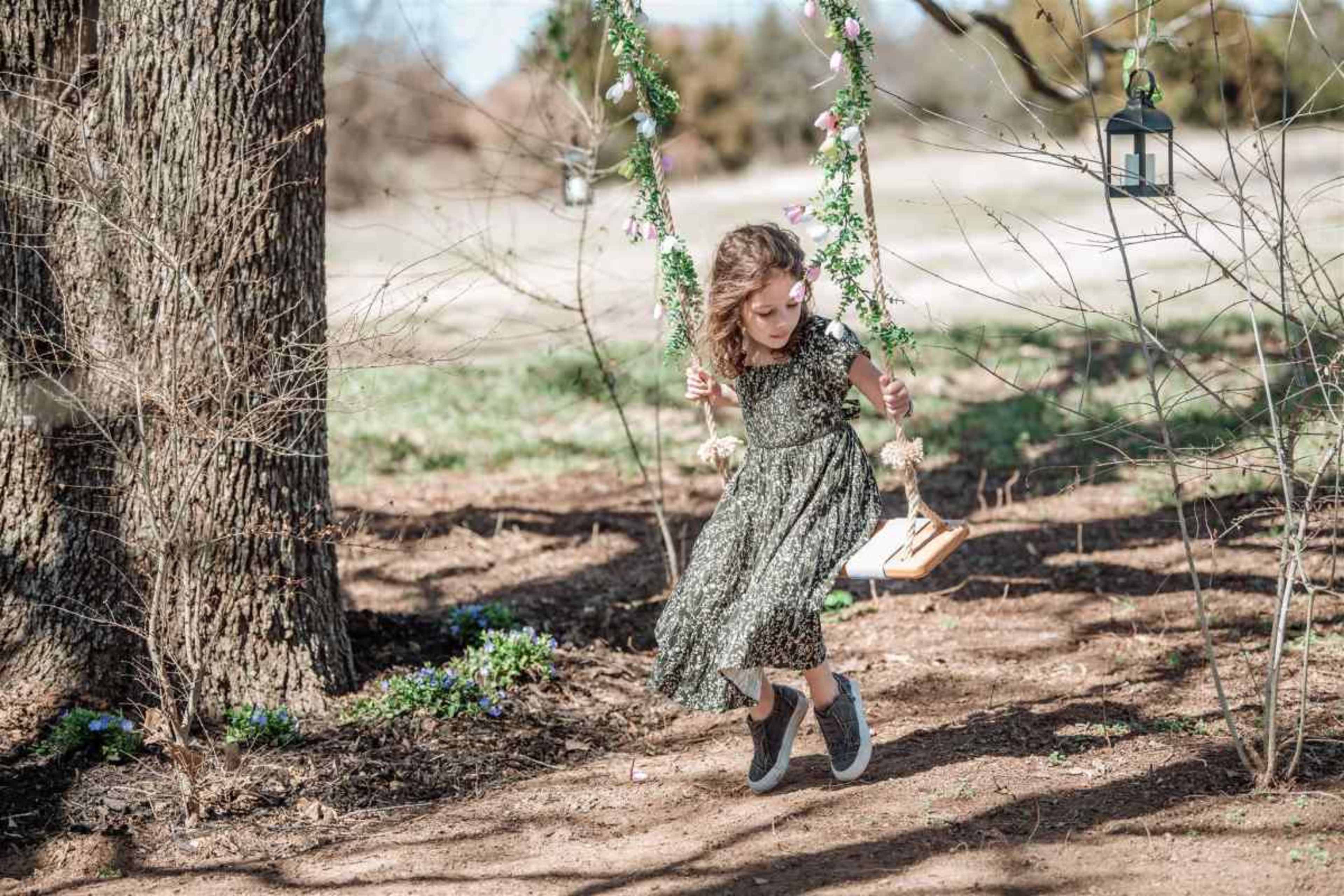 A young girl in a patterned dress swings on a wooden swing decorated with flowers, set in a grassy area near trees.