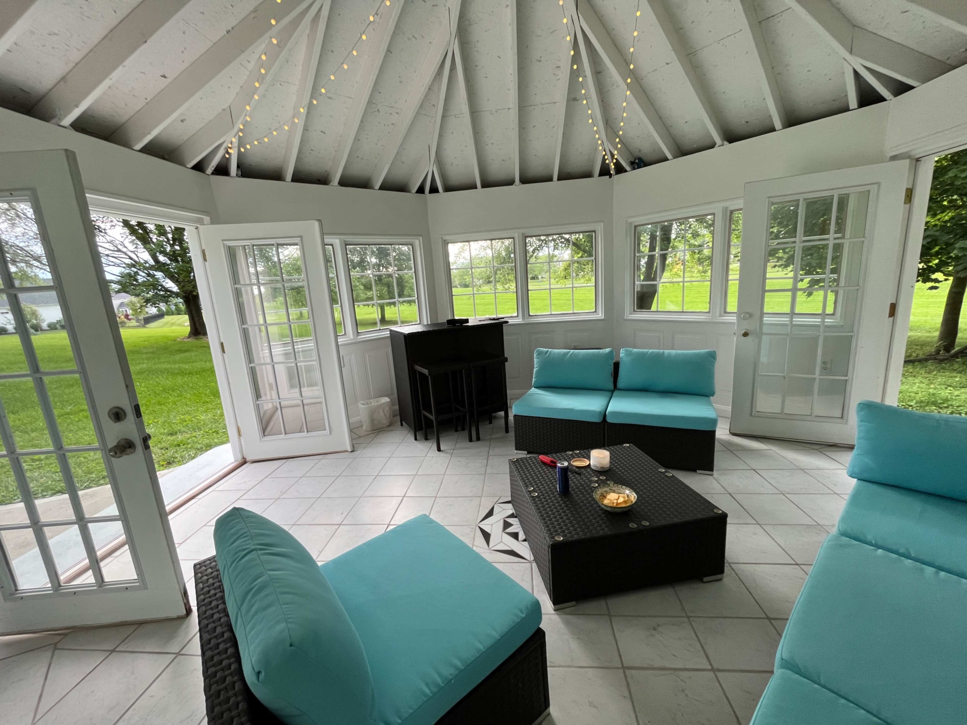 A gazebo with a tiled floor, featuring a seating area with turquoise cushions and a small table, surrounded by large windows and doors.