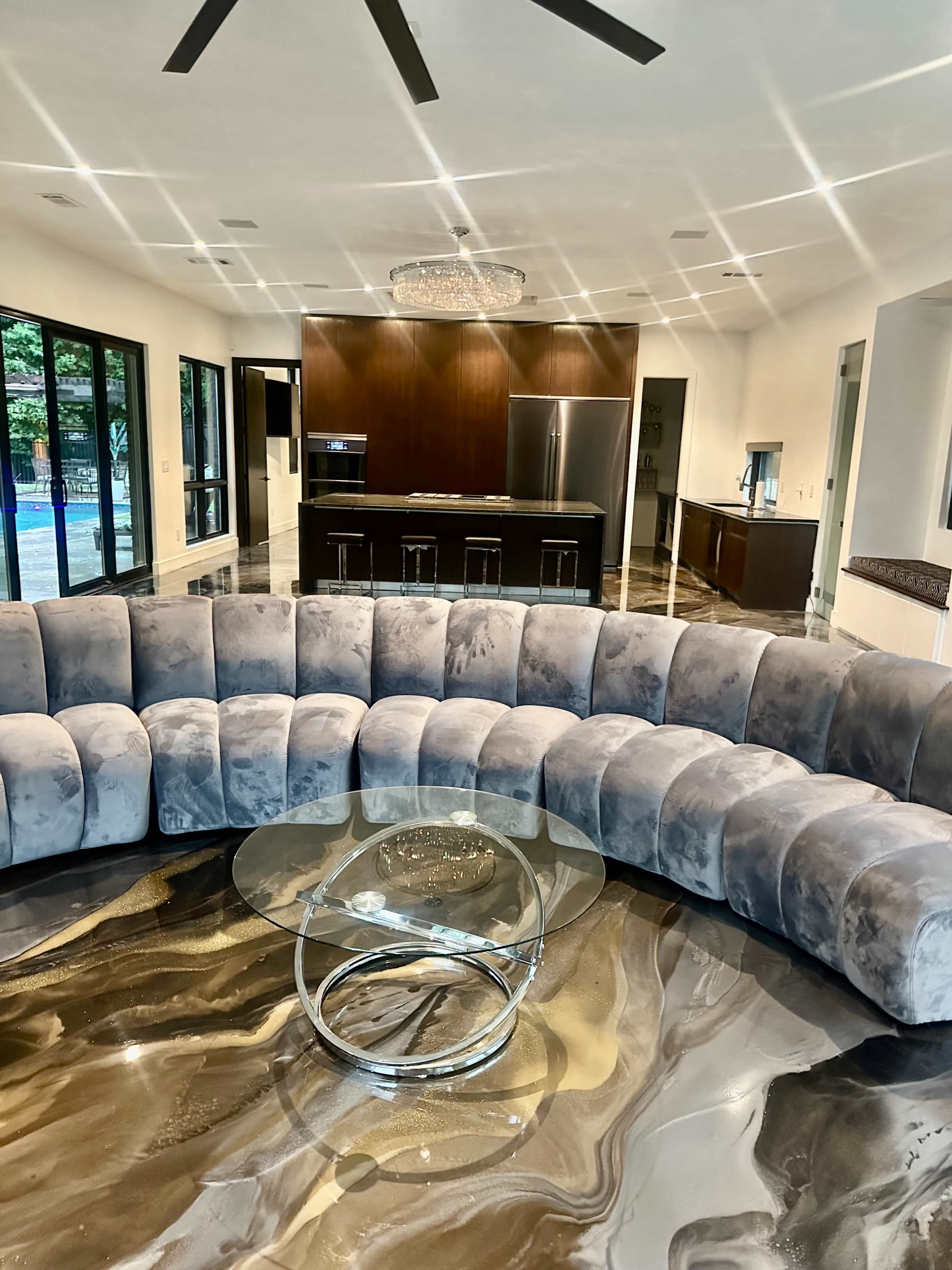 The image shows a curved gray sofa in a modern living room with a glass table, glossy patterned floor, and a kitchen area in the background.