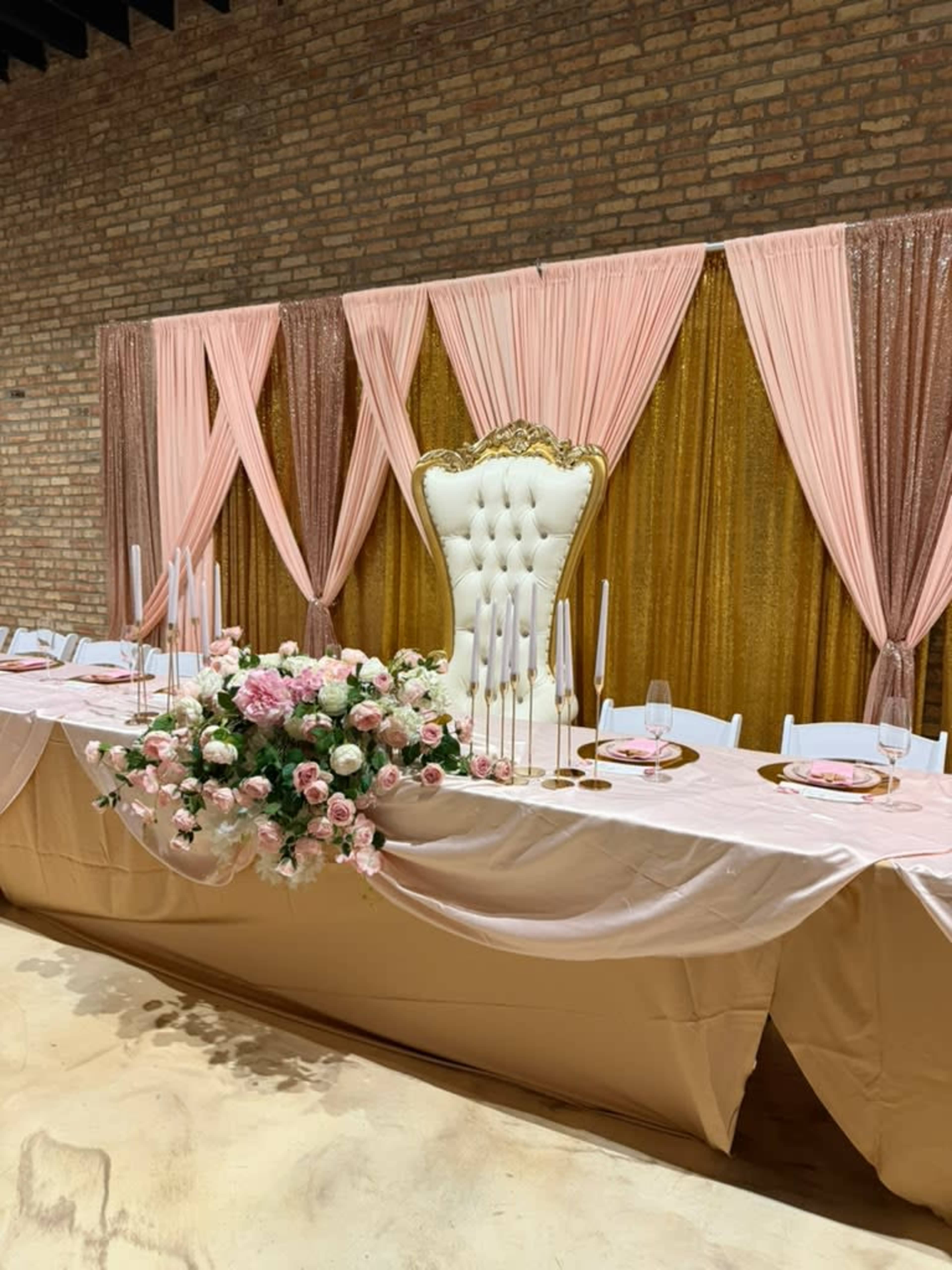 The image shows a decorated banquet table with a large white throne-like chair and pink drapery, set against a brick wall.