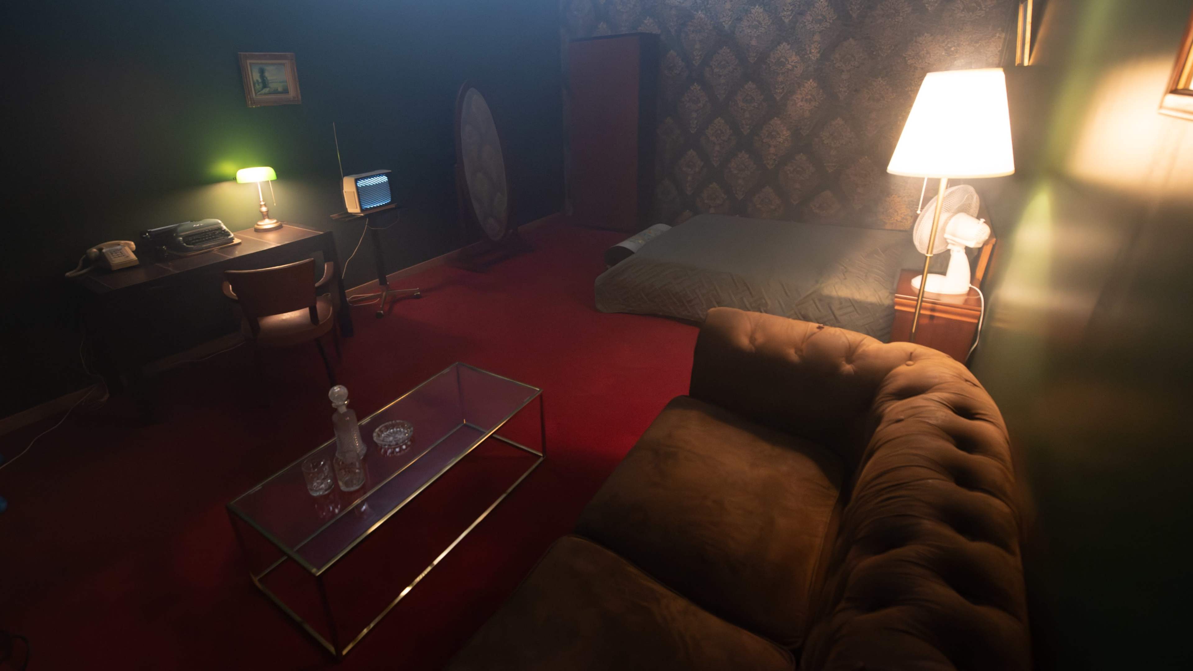 A dimly lit room with a bed, a vintage-style desk and chair, an old television, a lamp, and a glass coffee table with decanters.