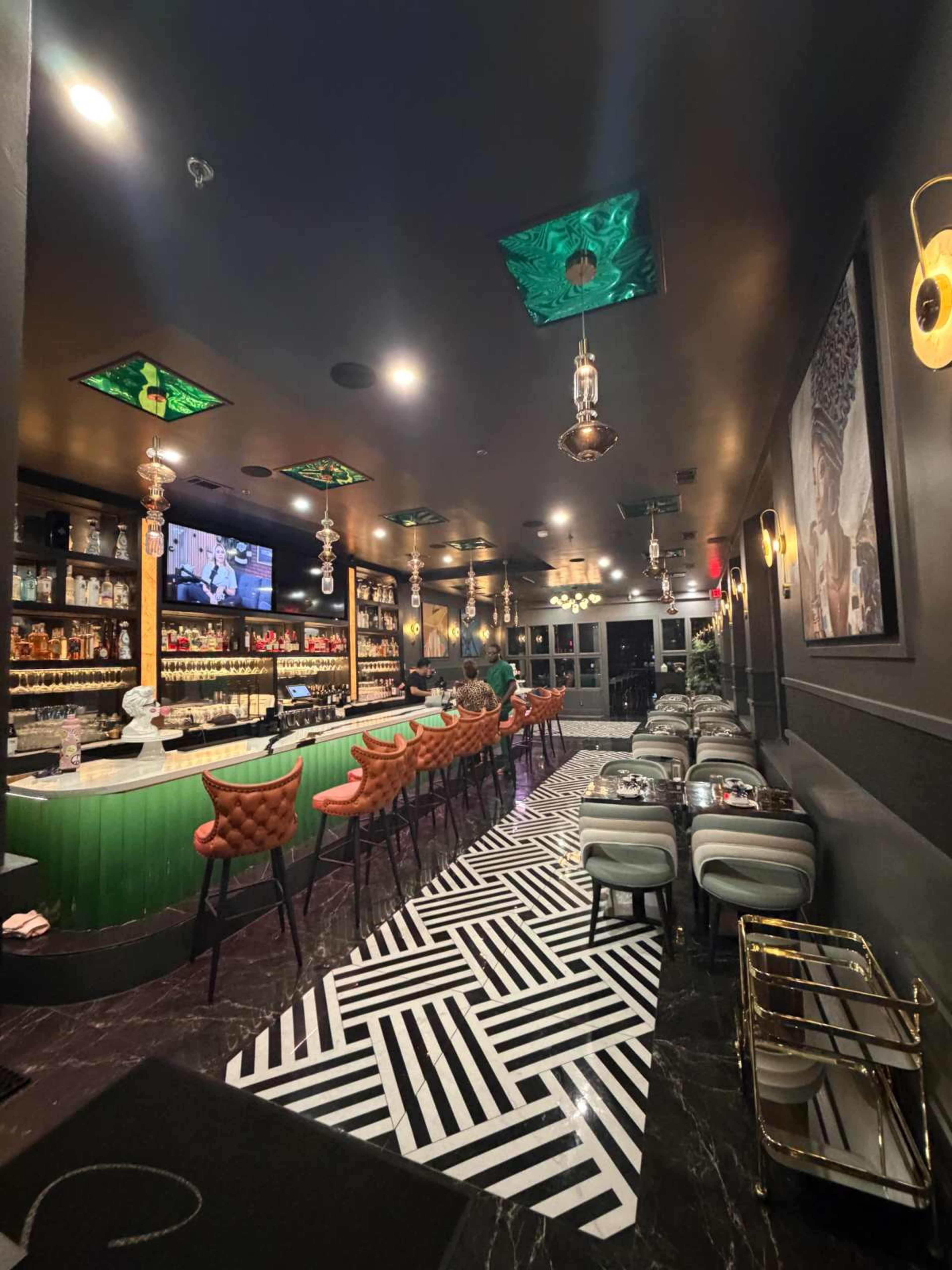 The image shows a modern bar with a green counter, stylish seating, and a patterned floor, illuminated by unique light fixtures.