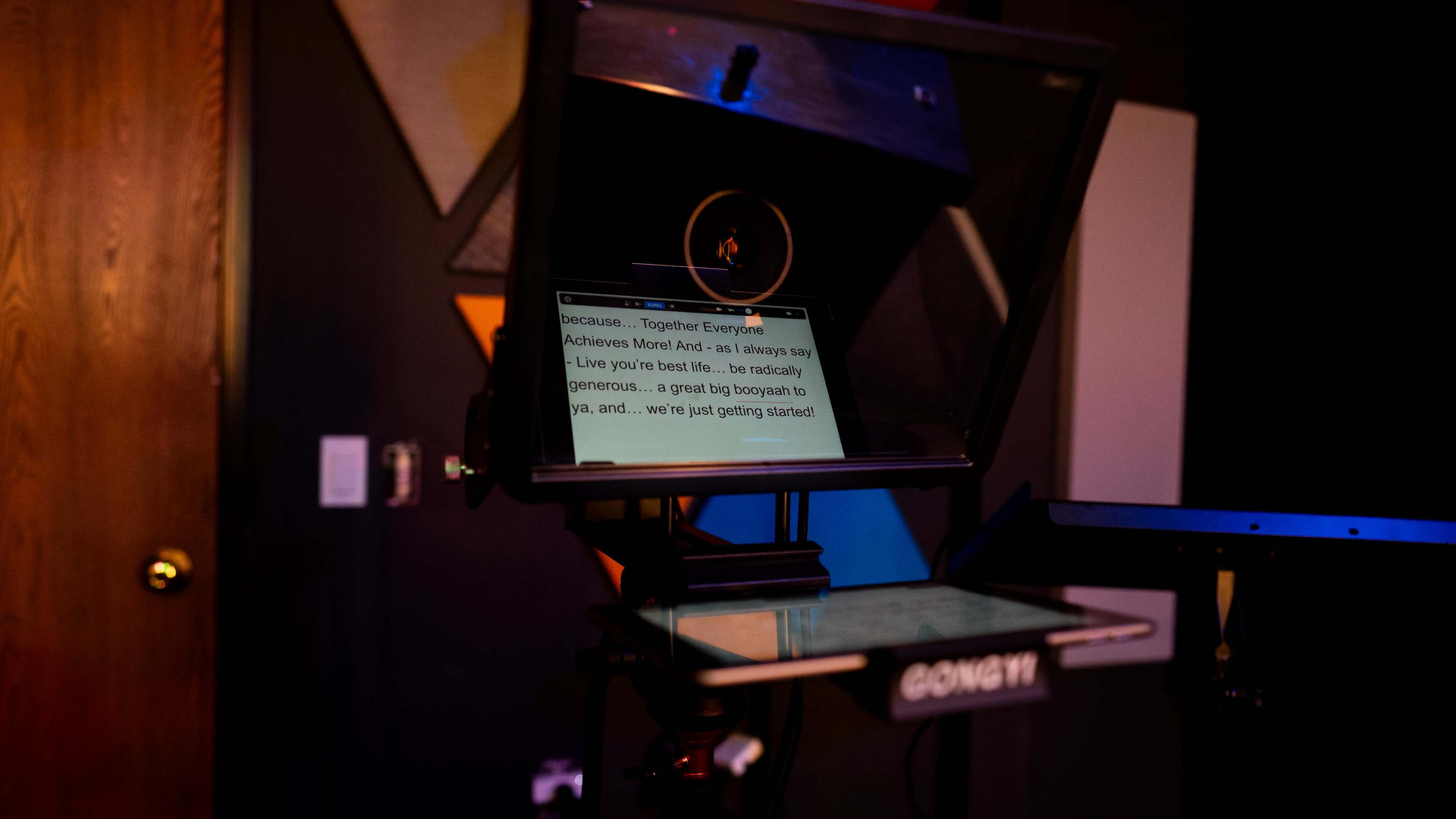 A teleprompter displays scripted text in a dimly lit room with colorful geometric wall decorations.