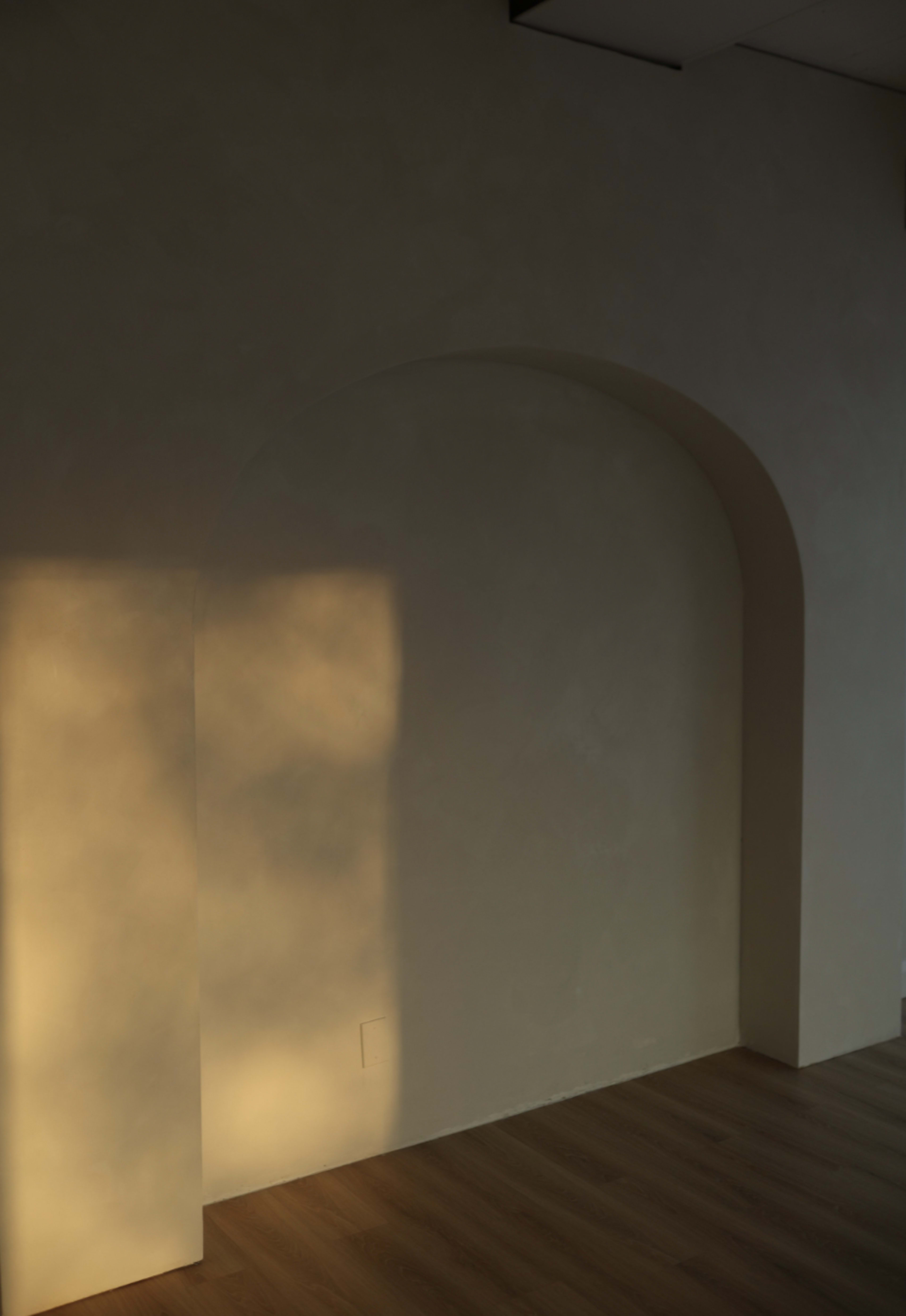 A minimalistic interior with a textured wall, an arched alcove, and sunlight casting a warm glow on the surface.