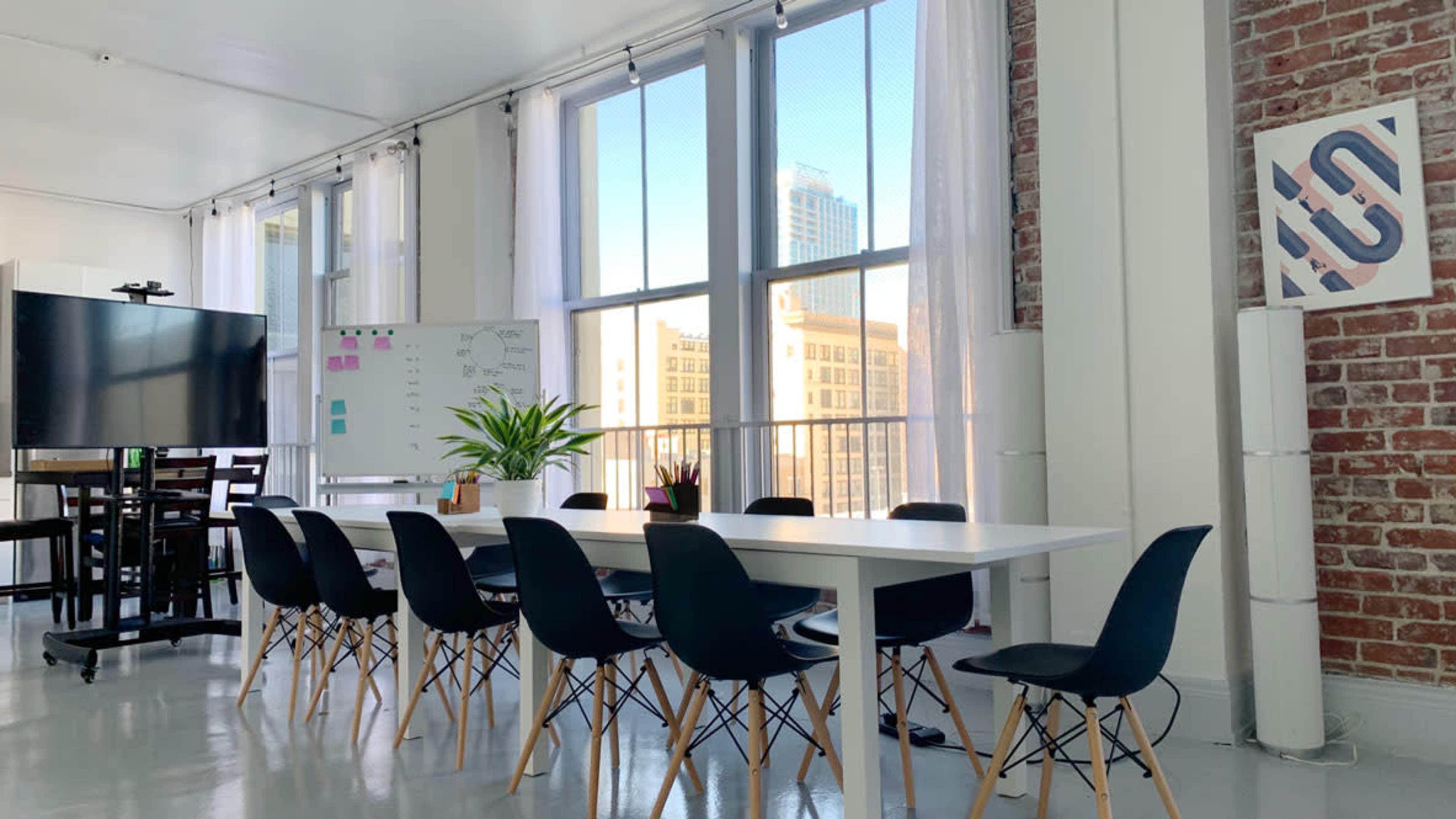 A modern meeting room with a long white table surrounded by black chairs, large windows providing natural light, and a digital screen on one side.