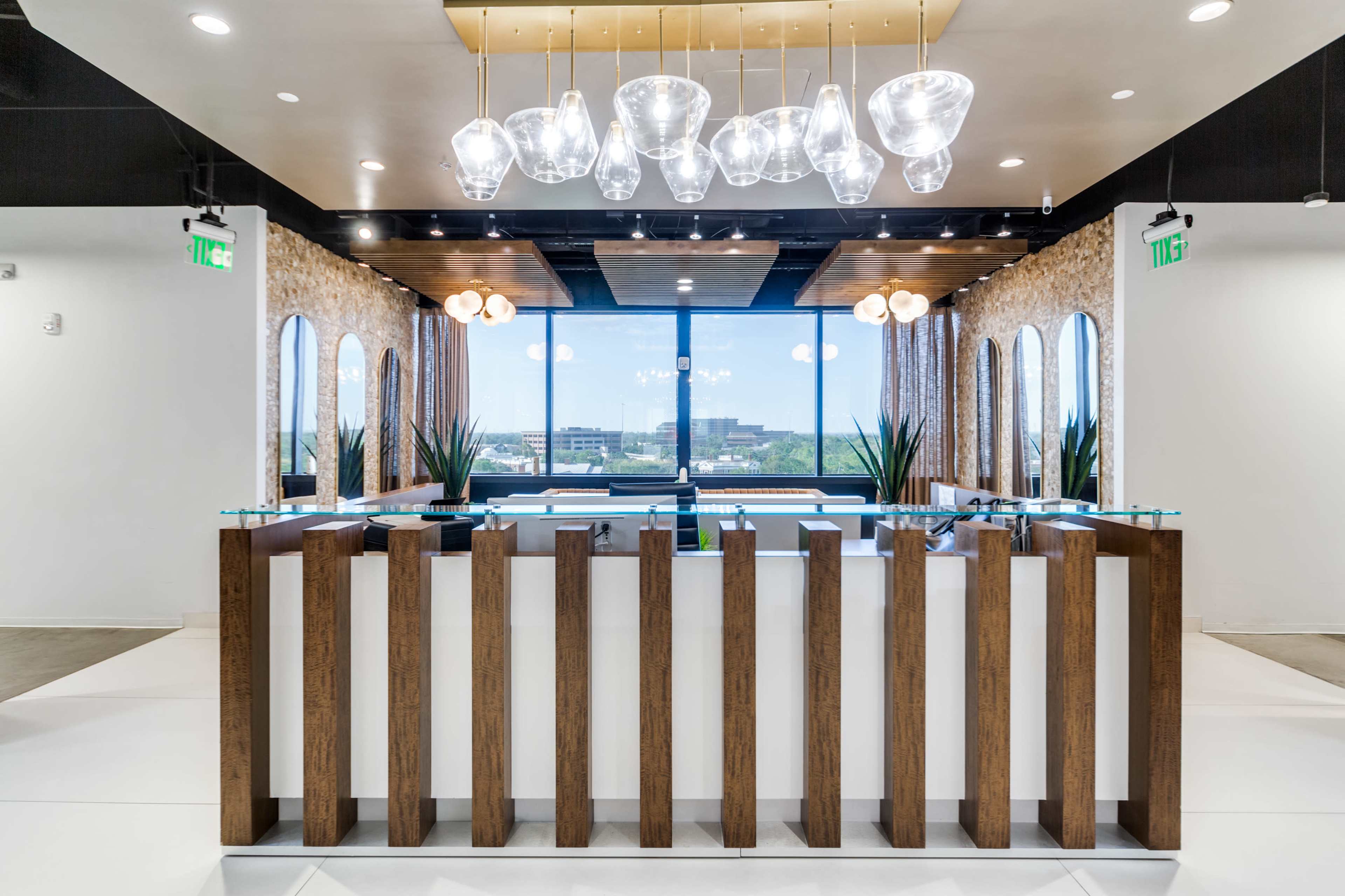 A modern reception area features a sleek wooden front desk with glass accents and large windows providing a view of the outside.