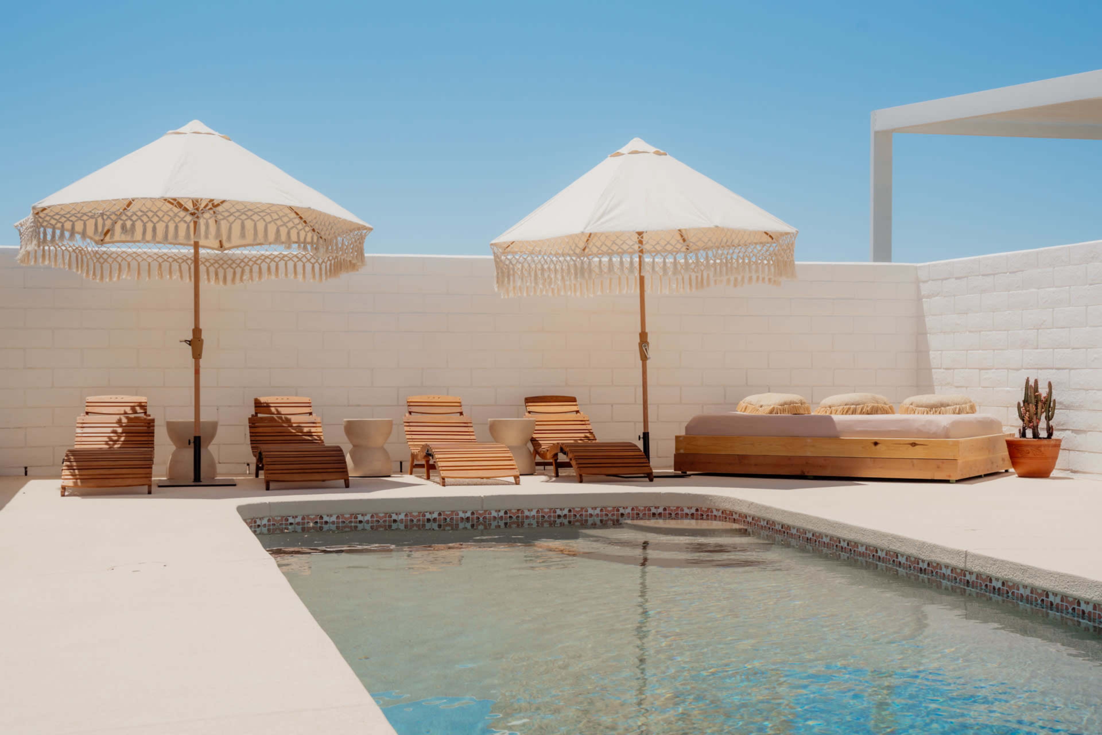 The image shows a poolside area featuring two large umbrellas, a wooden lounge, and several wooden lounge chairs beside a clear blue swimming pool.