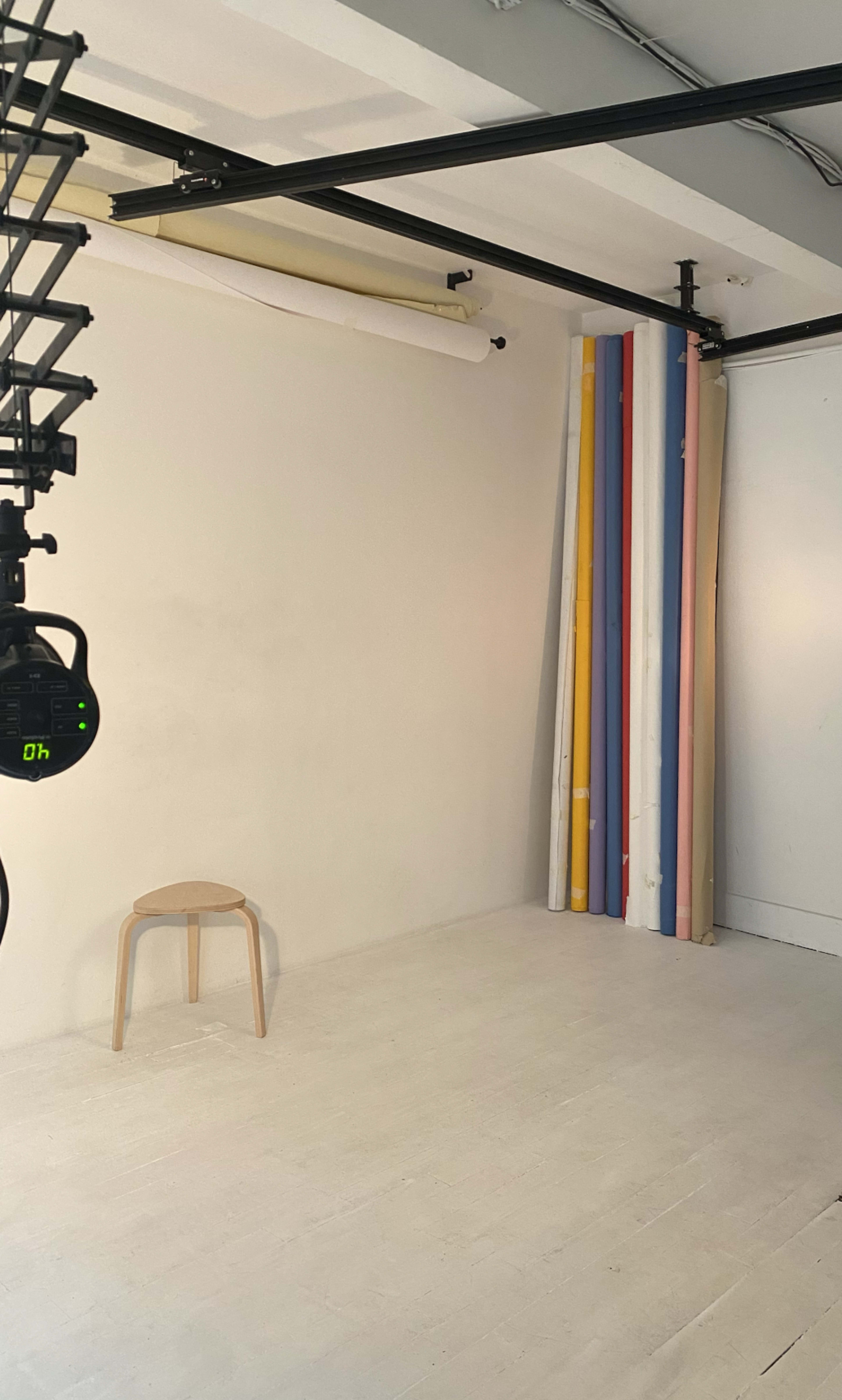 The image shows a minimalist photography studio with a wooden stool in the foreground and several colorful backdrop rolls leaning against the wall.