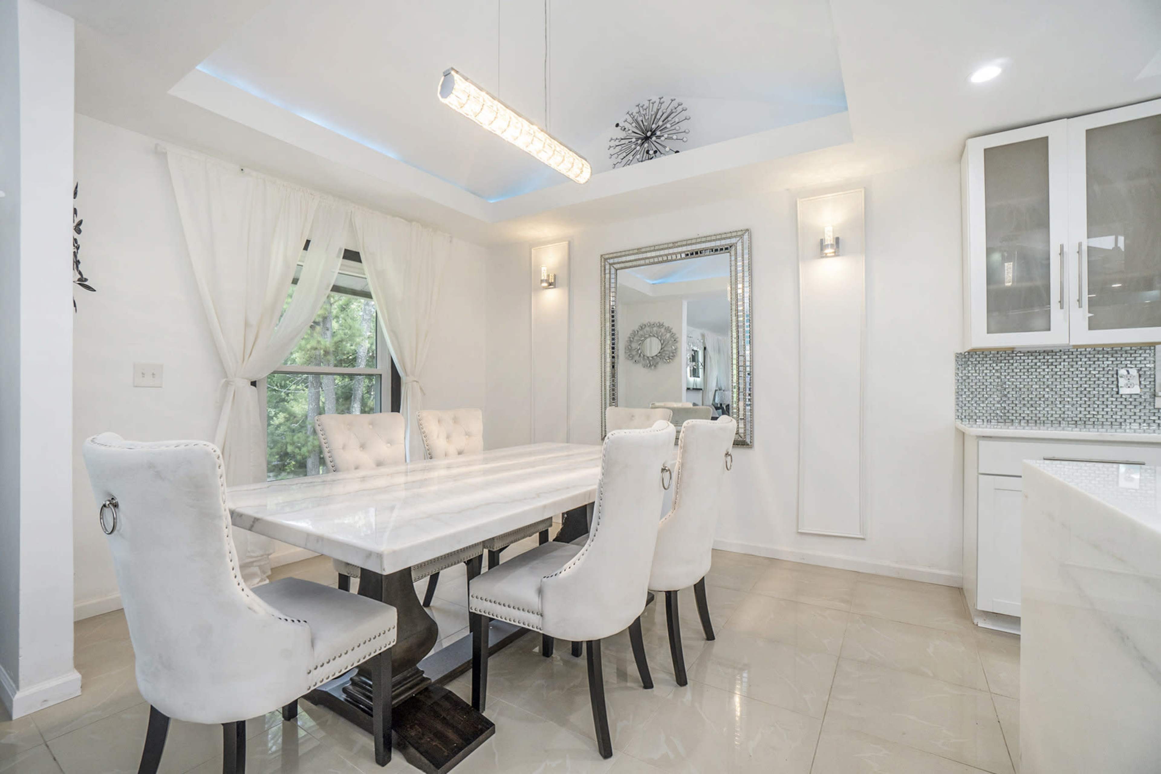 A modern dining area features a long marble table surrounded by upholstered chairs, with large windows allowing natural light to enter.