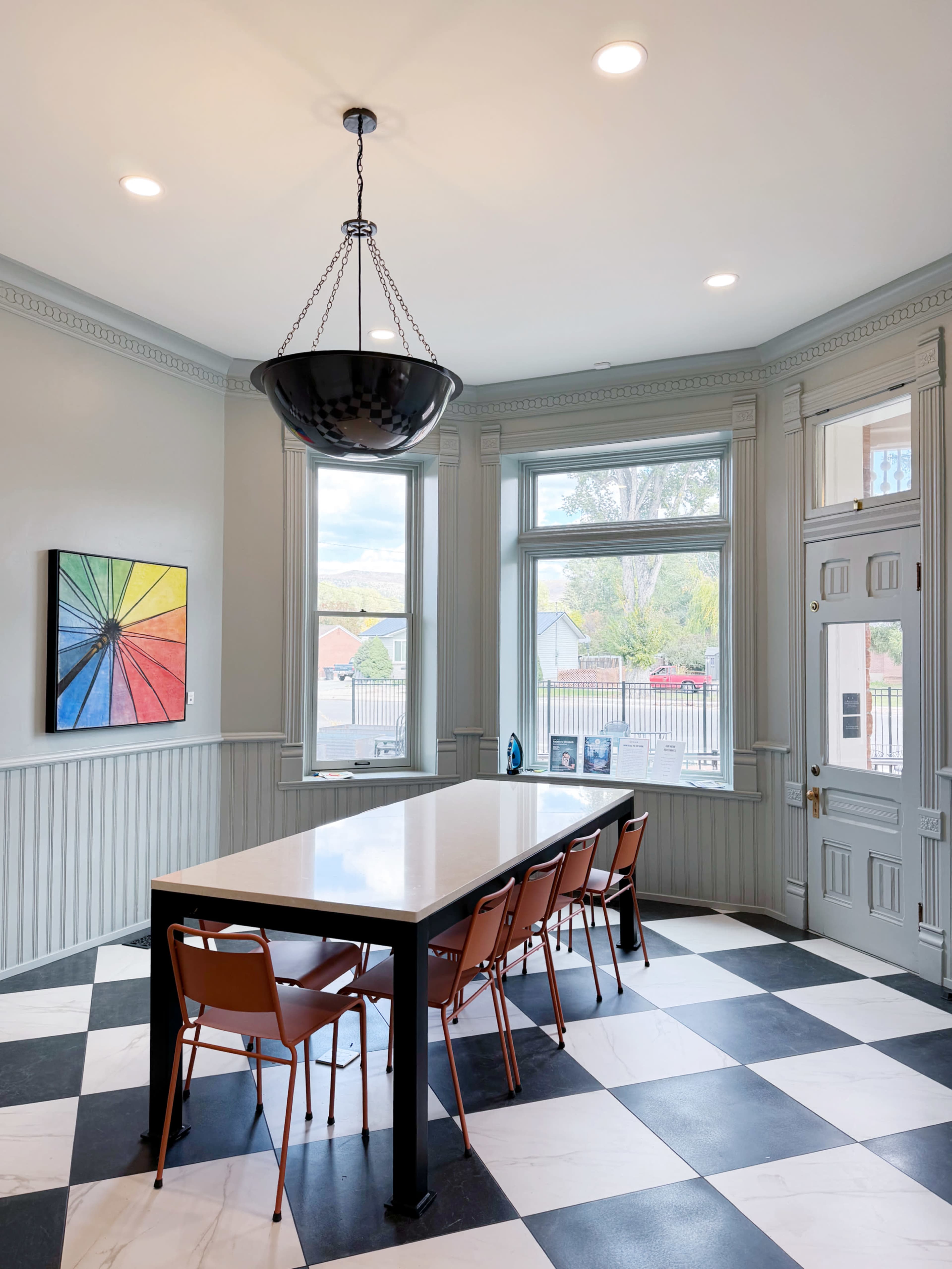 A modern dining area features a large rectangular table surrounded by six chairs, with a colorful abstract painting on the wall and large windows allowing natural light to enter.