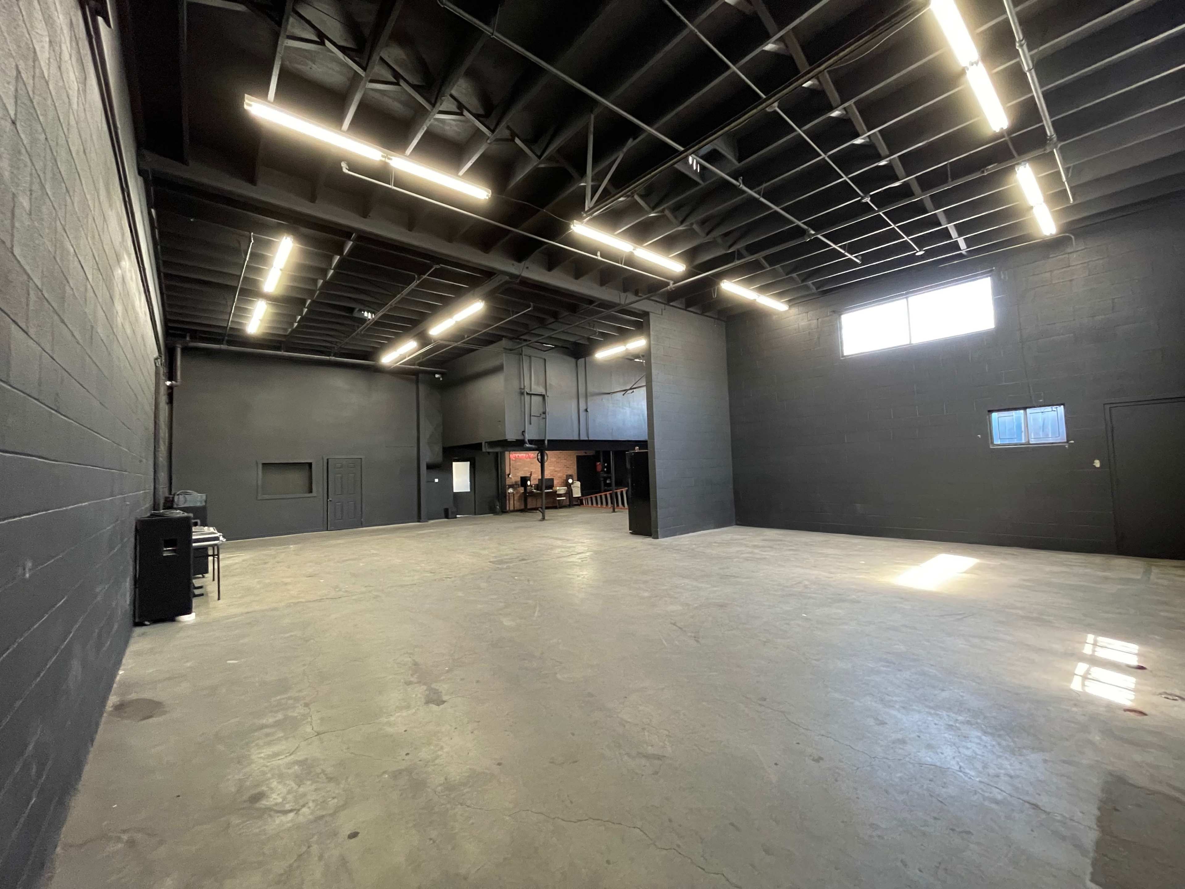 A spacious, empty industrial room with black walls and a concrete floor, featuring overhead lighting and a few windows.