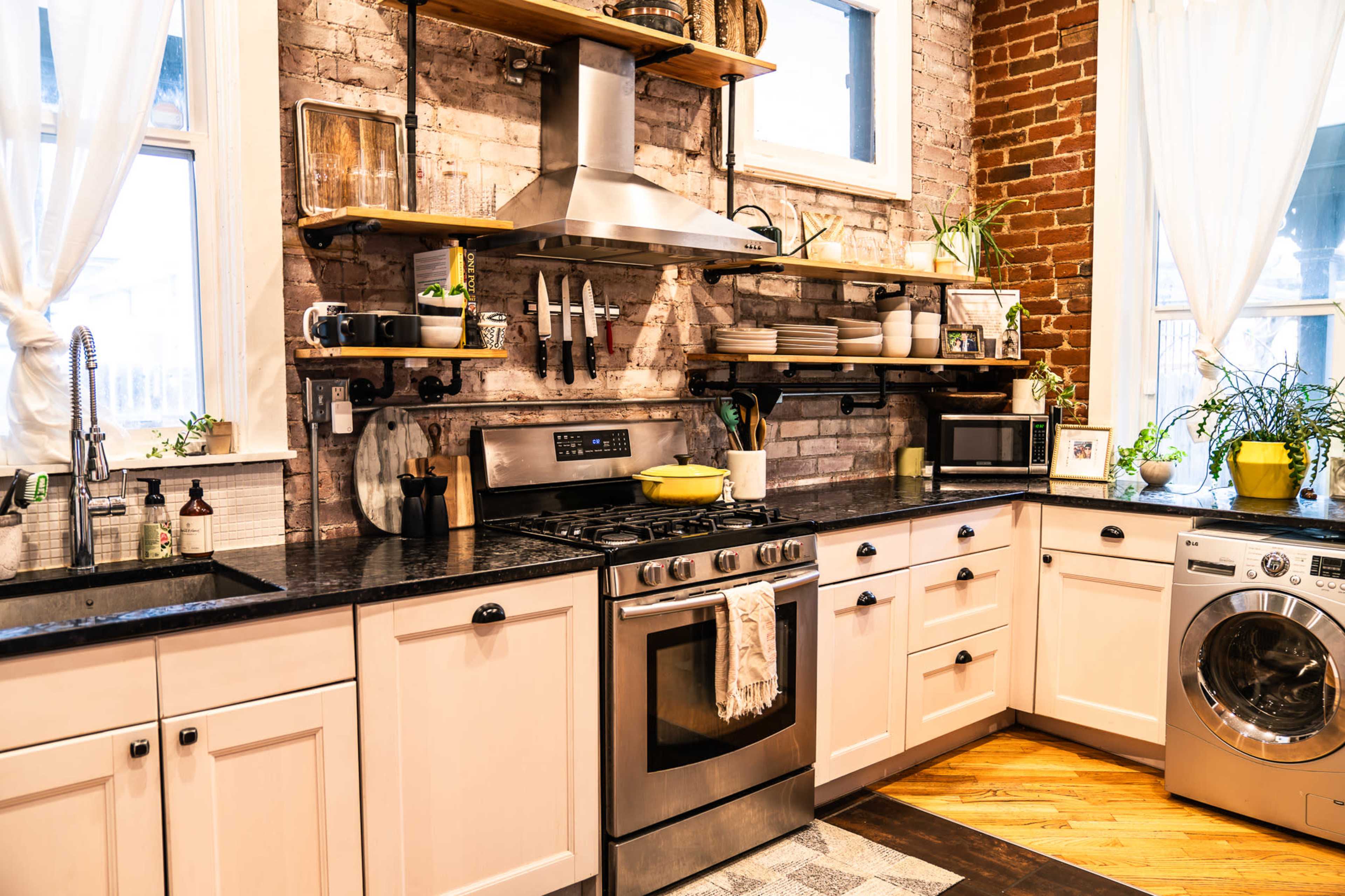 The kitchen features a stainless steel stove, open shelves with various kitchenware, and a washing machine, all within a brick-walled interior.