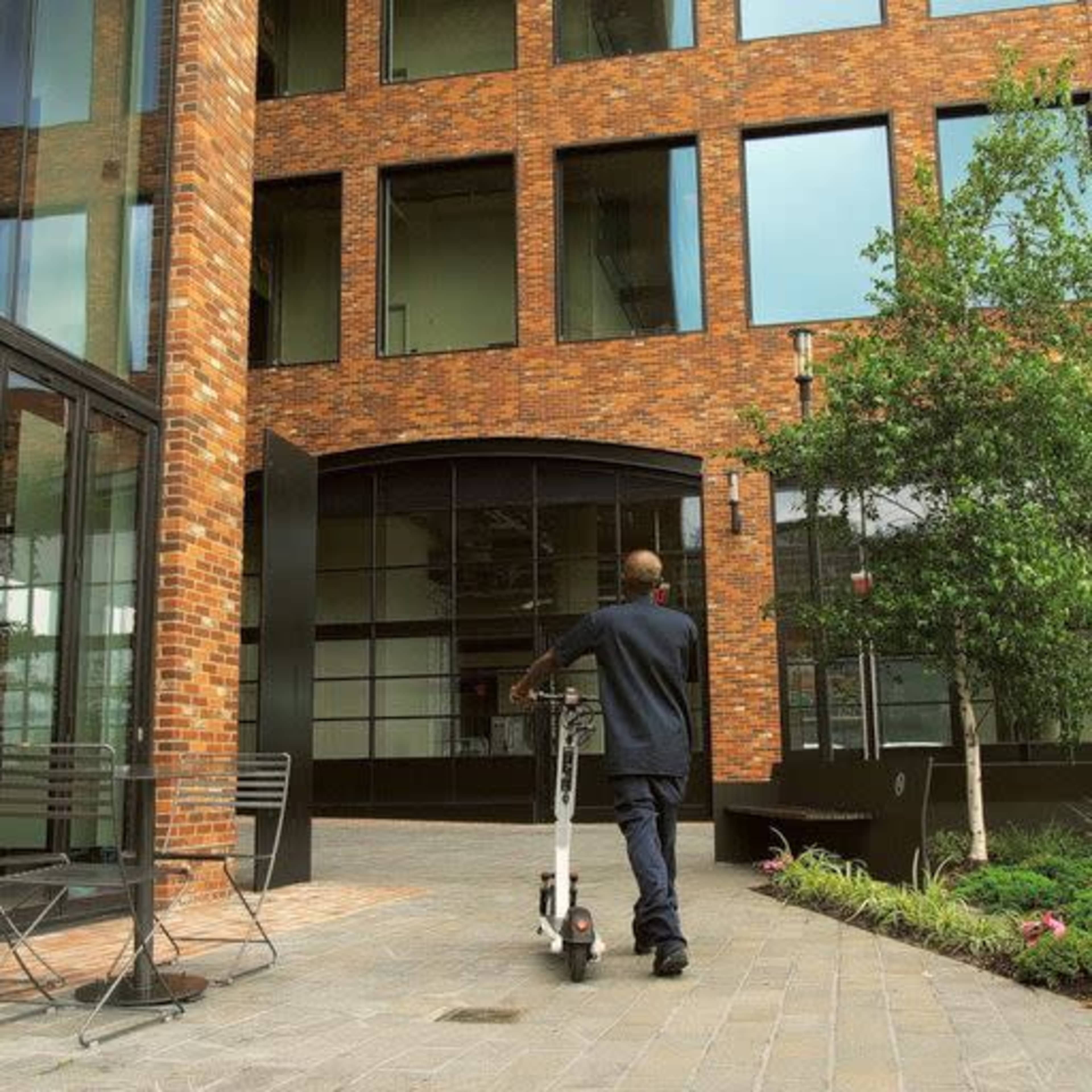 A person in a dark outfit rides an electric scooter past a brick building with large windows.