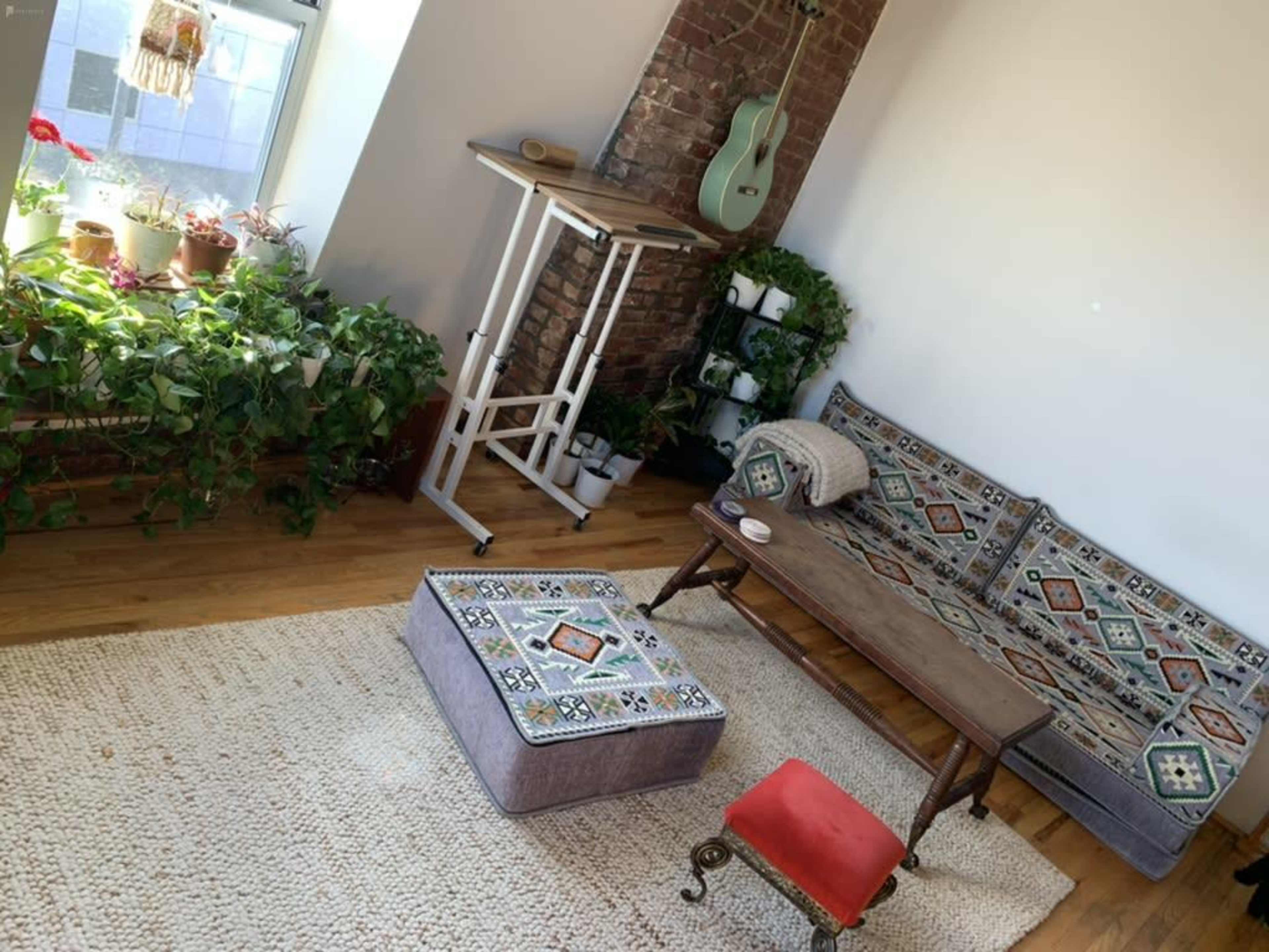 A bright room with a brick wall, a couch adorned with patterned cushions, a wooden table beside a green guitar, and a small red ottoman on a woven rug.