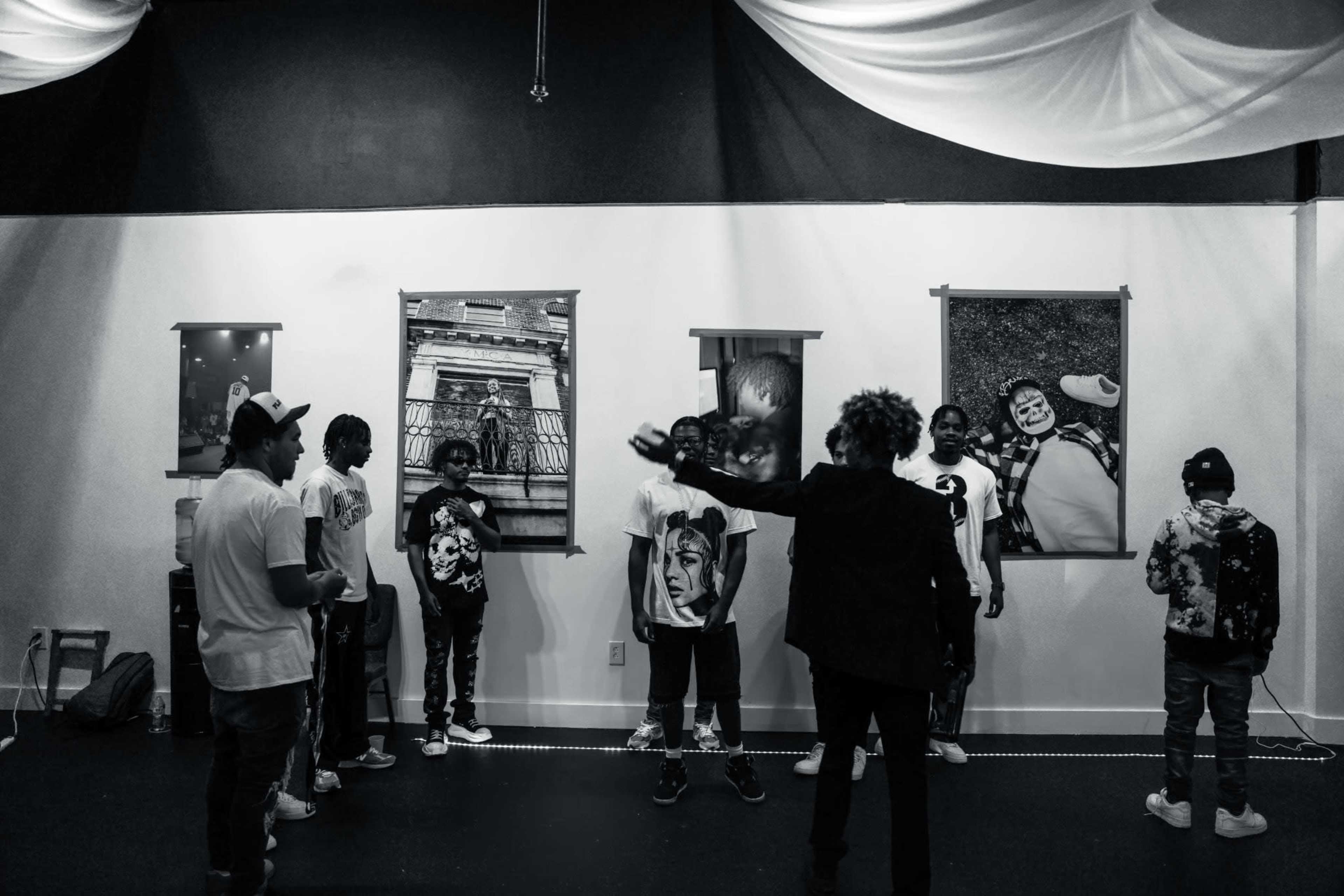 A group of people stands in a gallery space with framed artwork on the walls, while another person gestures toward them.