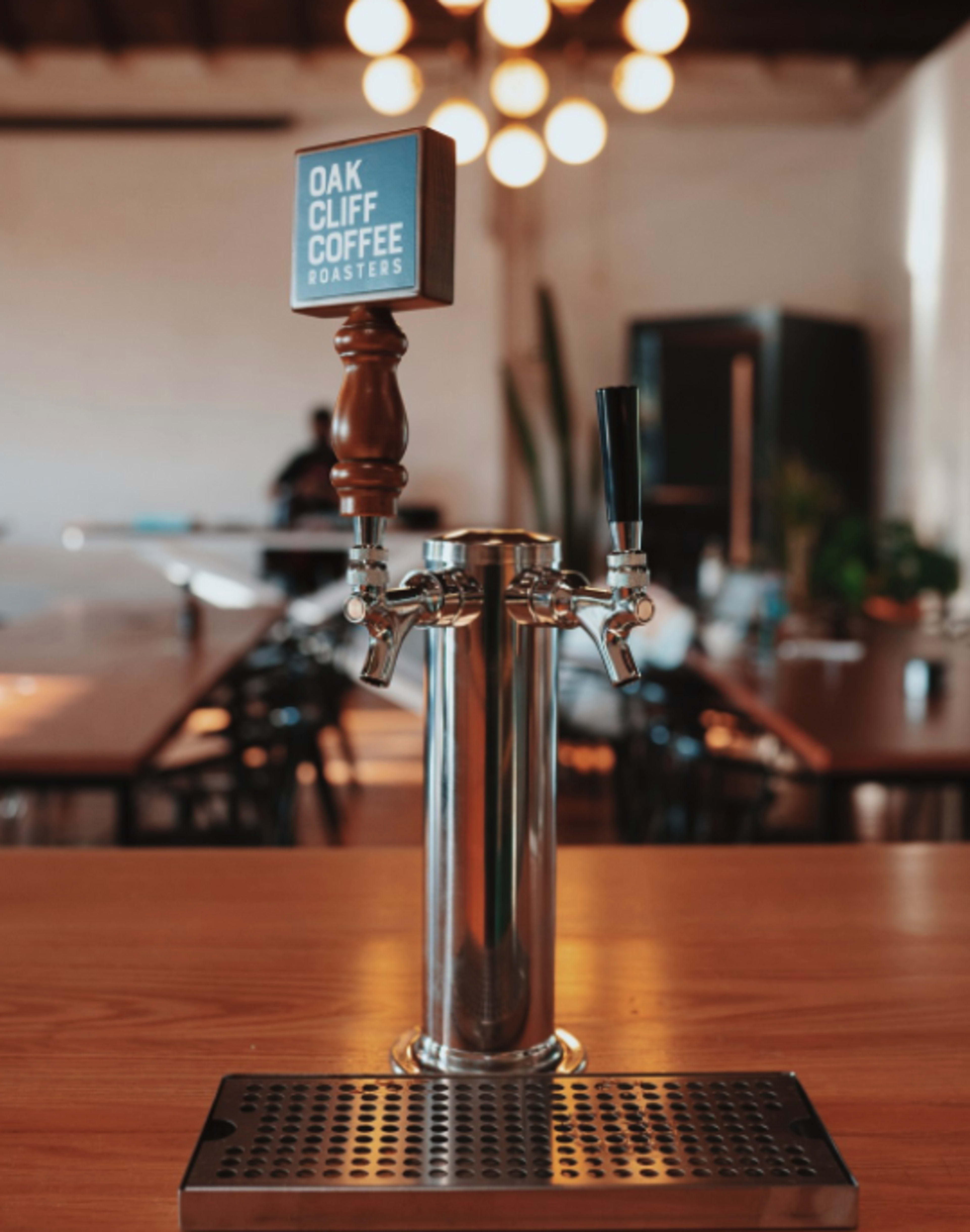 A polished metal coffee tap with a wooden handle and a sign reading "Oak Cliff Coffee Roasters" stands on a wooden table in a well-lit room with hanging lights and plants in the background.