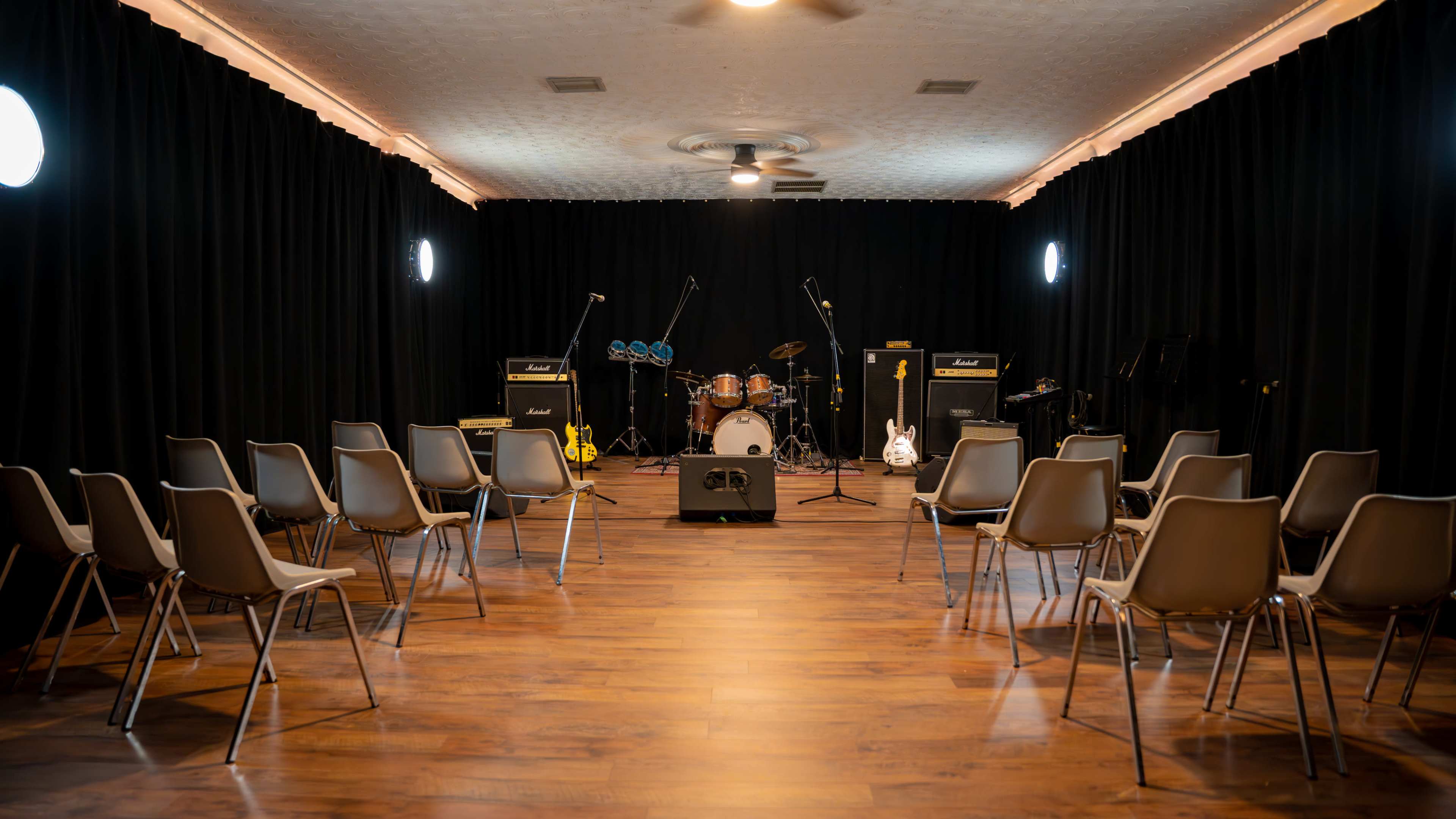 A small music performance space with rows of chairs facing a drum set and amplifiers against a black curtain backdrop.