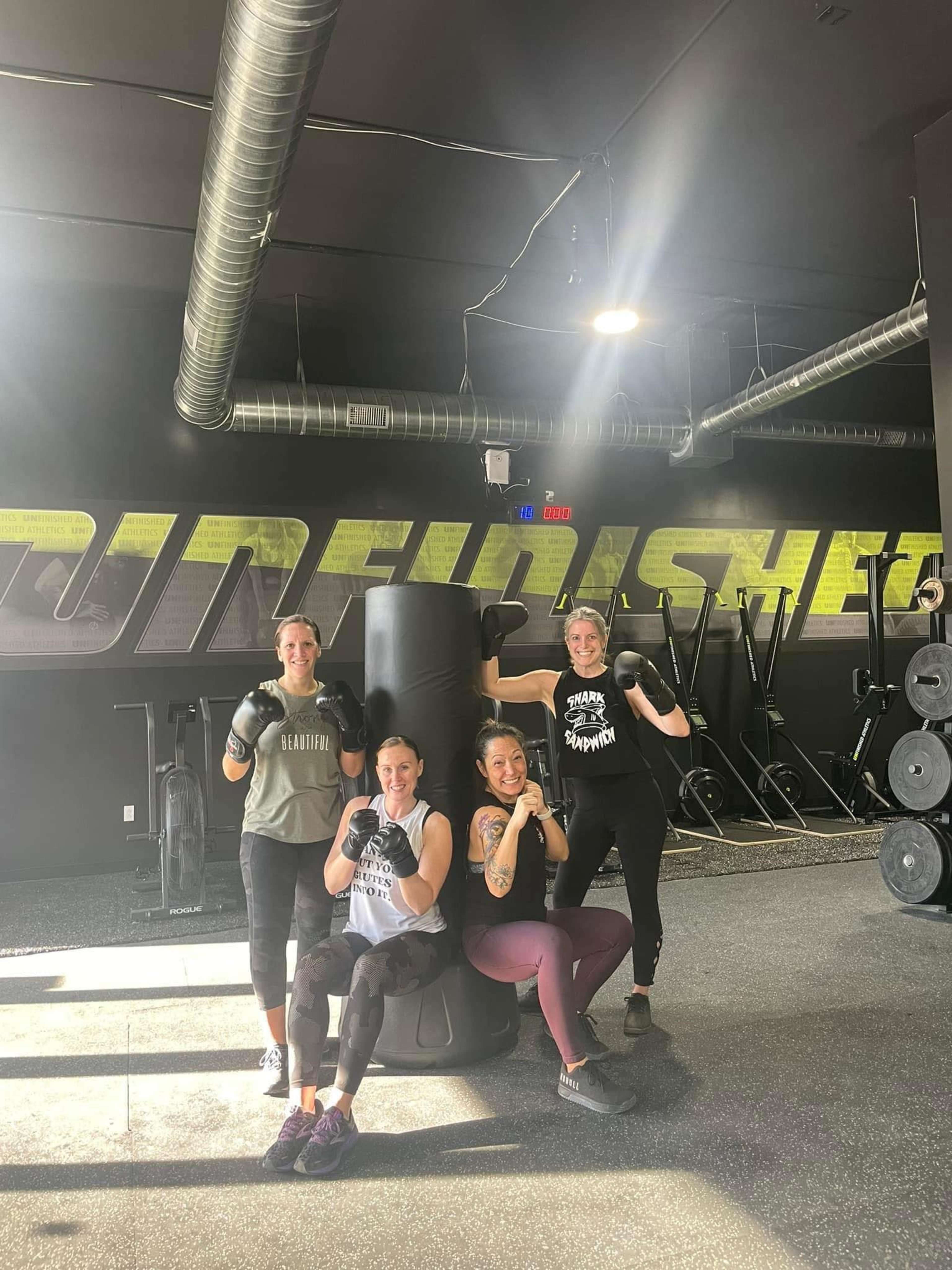 Four women pose with boxing gloves in a gym setting, standing in front of a punching bag and surrounded by fitness equipment.