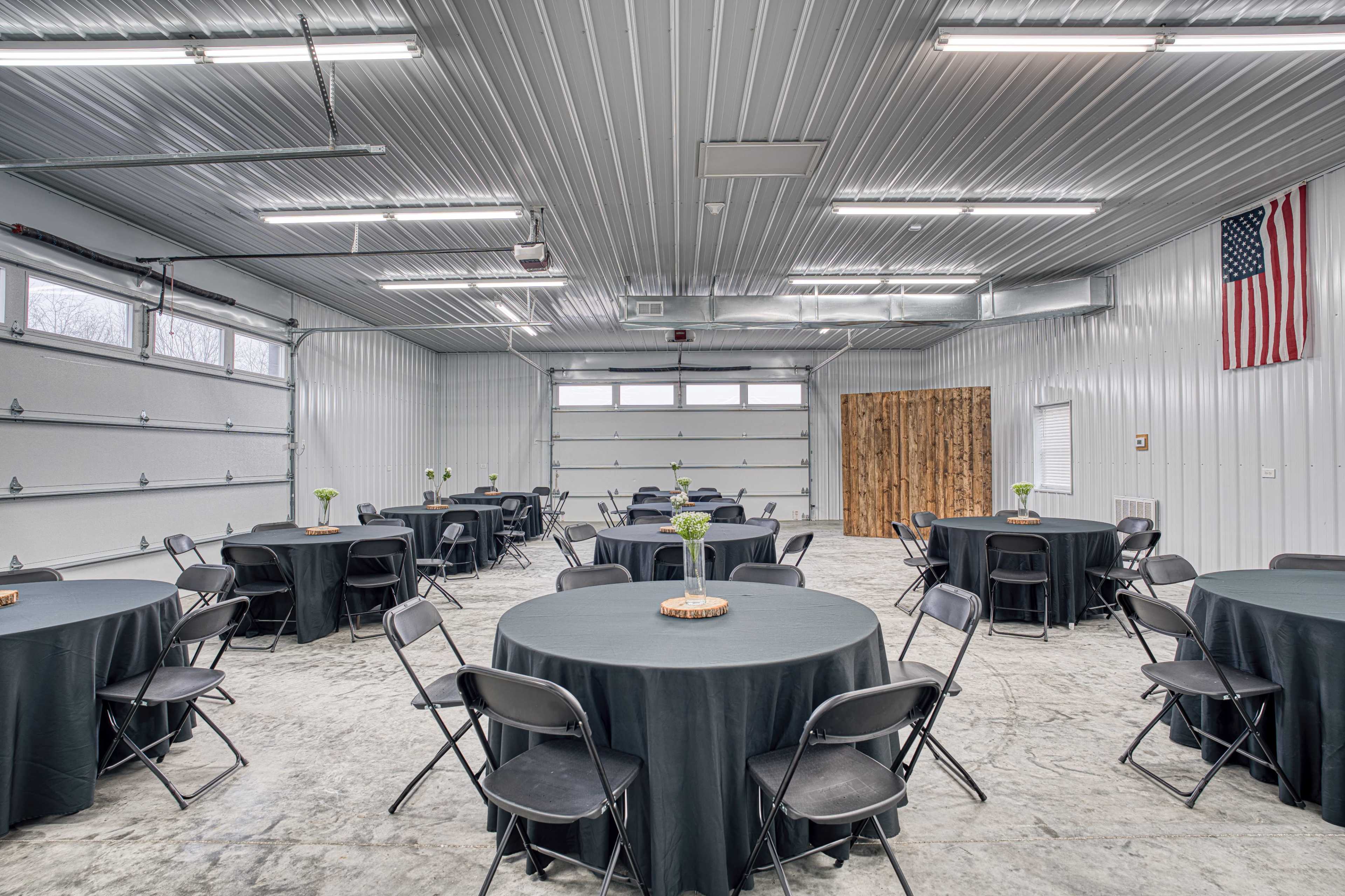 The image shows a spacious indoor venue with circular tables covered in black cloth, arranged under a metal roof, and large garage doors on one side.