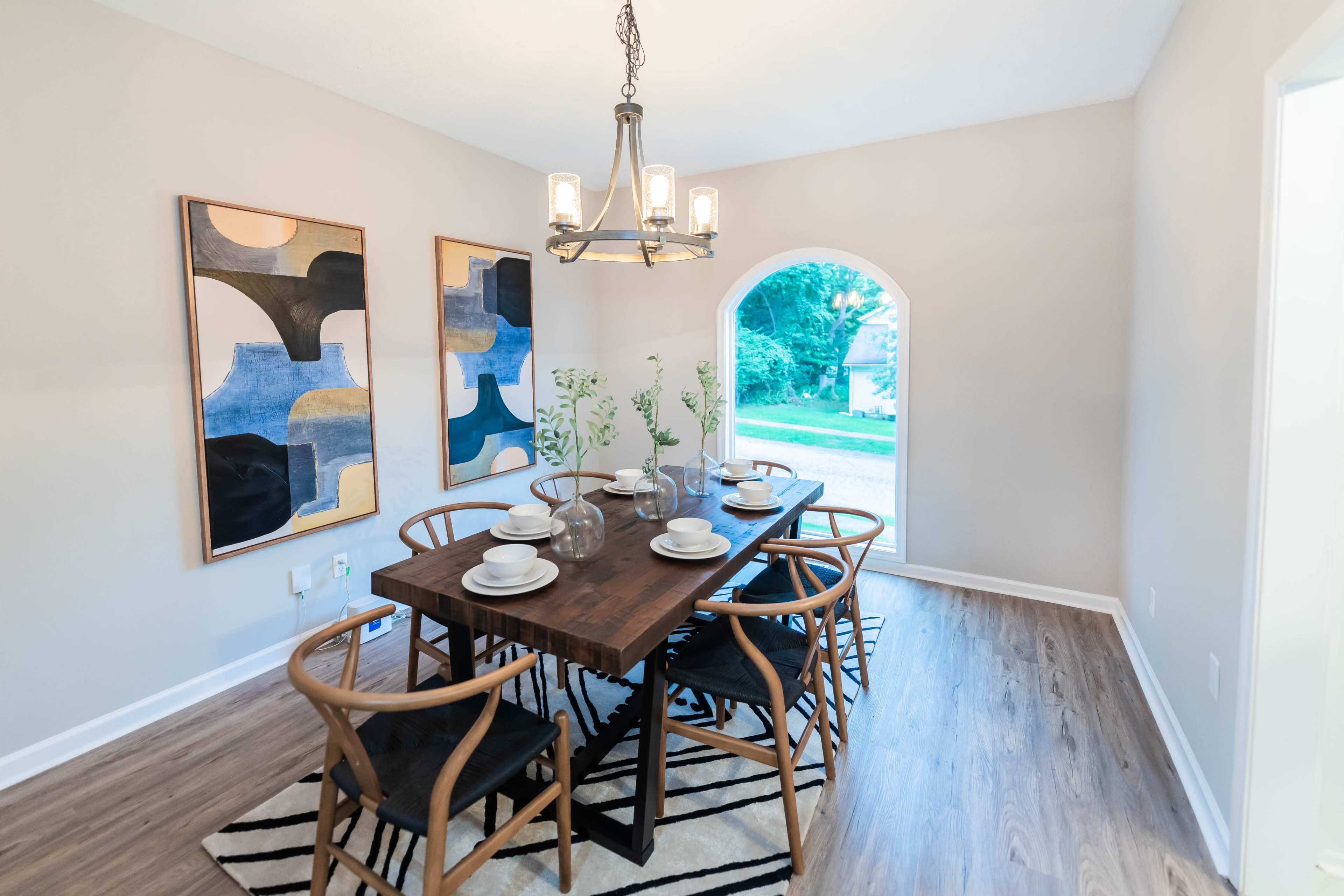 A dining room features a wooden table set for six, surrounded by curved chairs, with two abstract paintings on the walls and an arched doorway leading to an outdoor view.