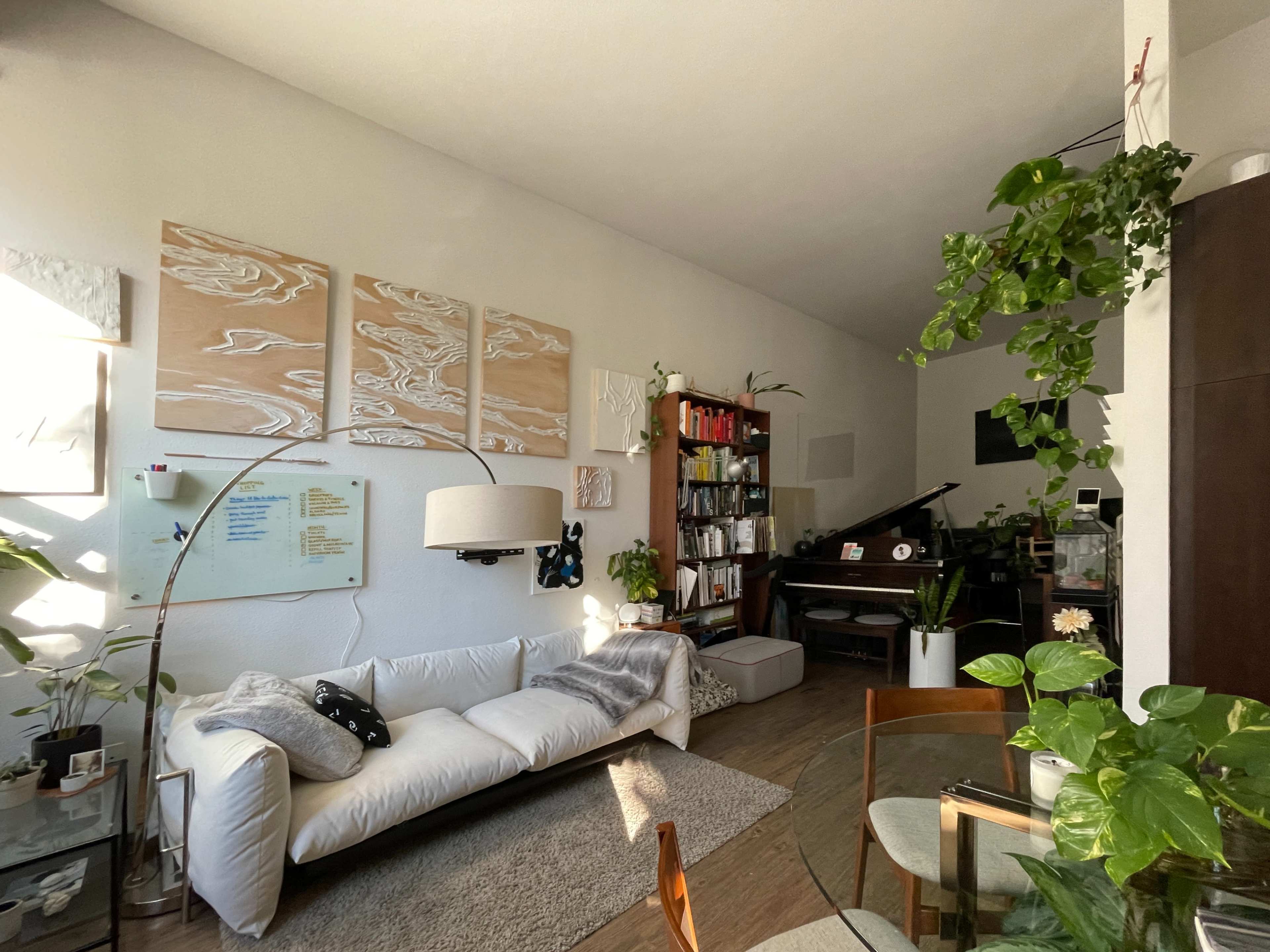 The image shows a cozy living room with a white sofa, bookshelf, piano, and various plants arranged around the space.