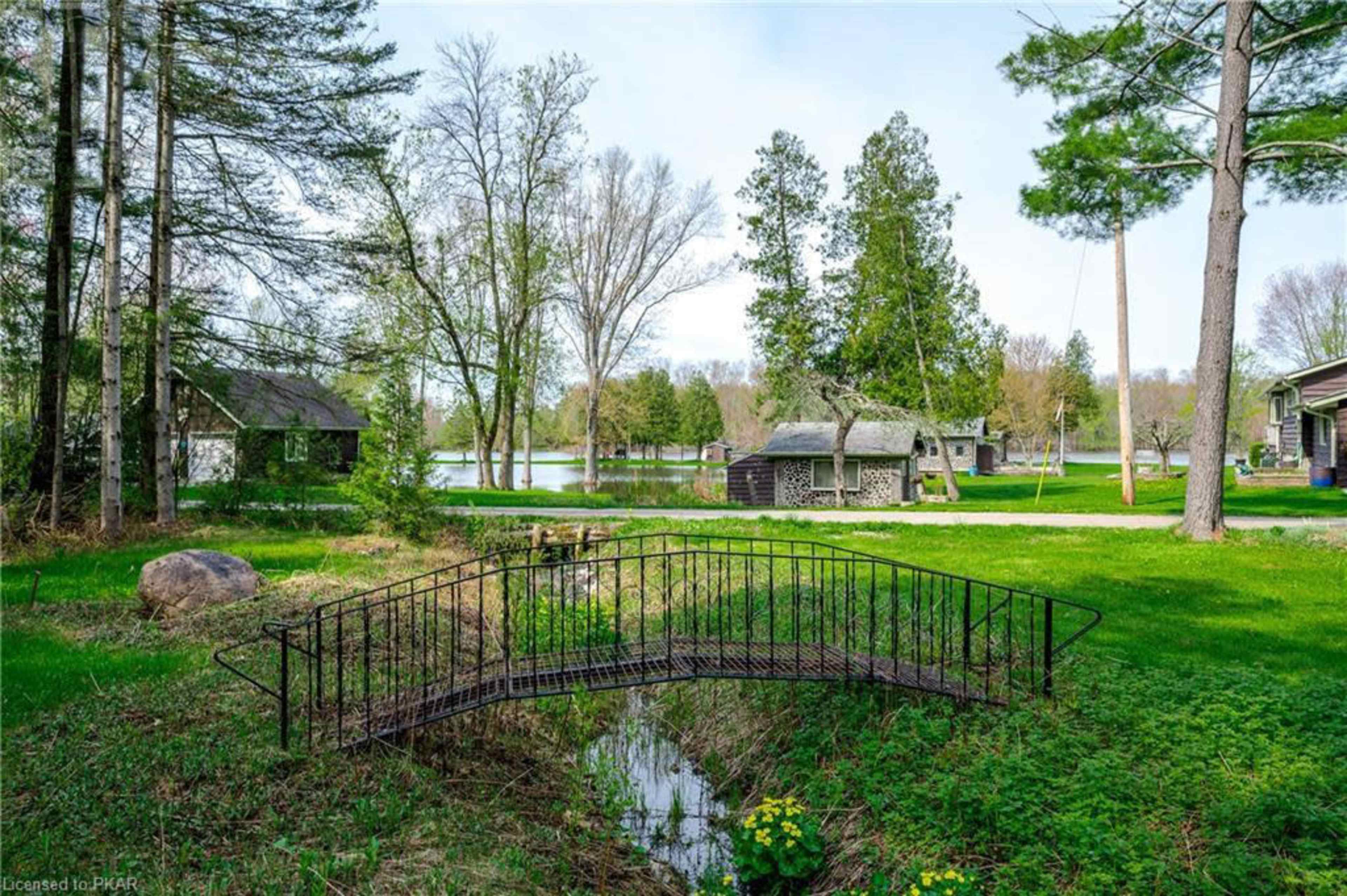 A metal bridge spans a small stream in a grassy area near several houses and trees by a lake.