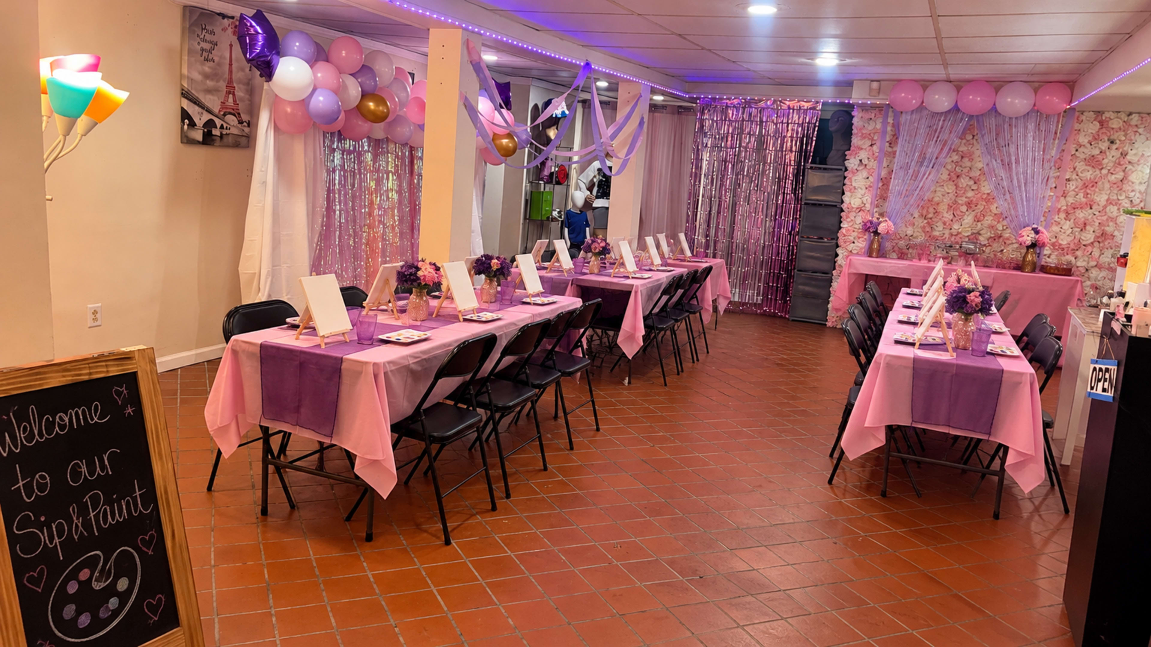 The image shows a decorated event space with rows of tables set up for a paint and sip party, featuring pink tablecloths, floral decorations, and colorful balloons.