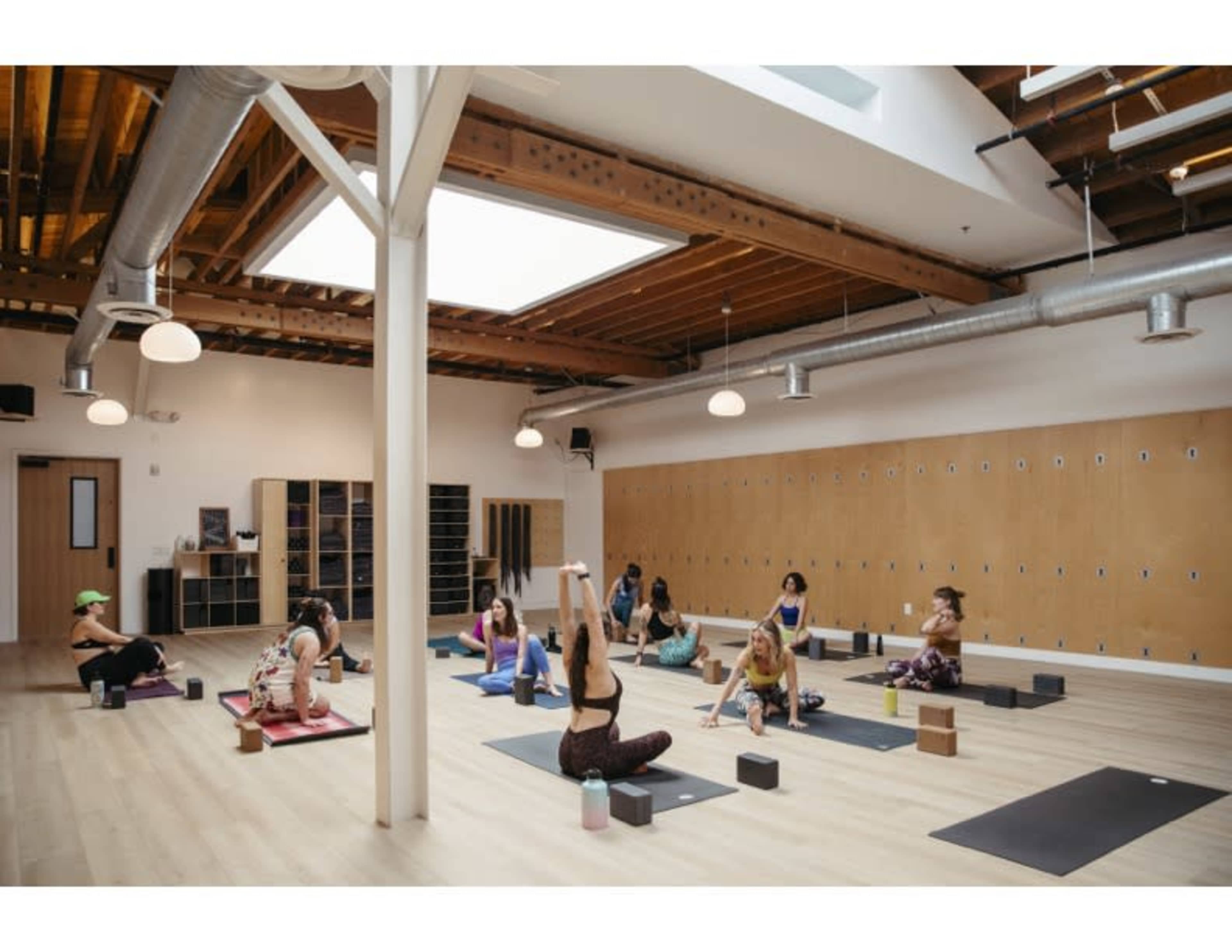 A group of individuals practices yoga in a spacious studio with wooden floors and large windows.