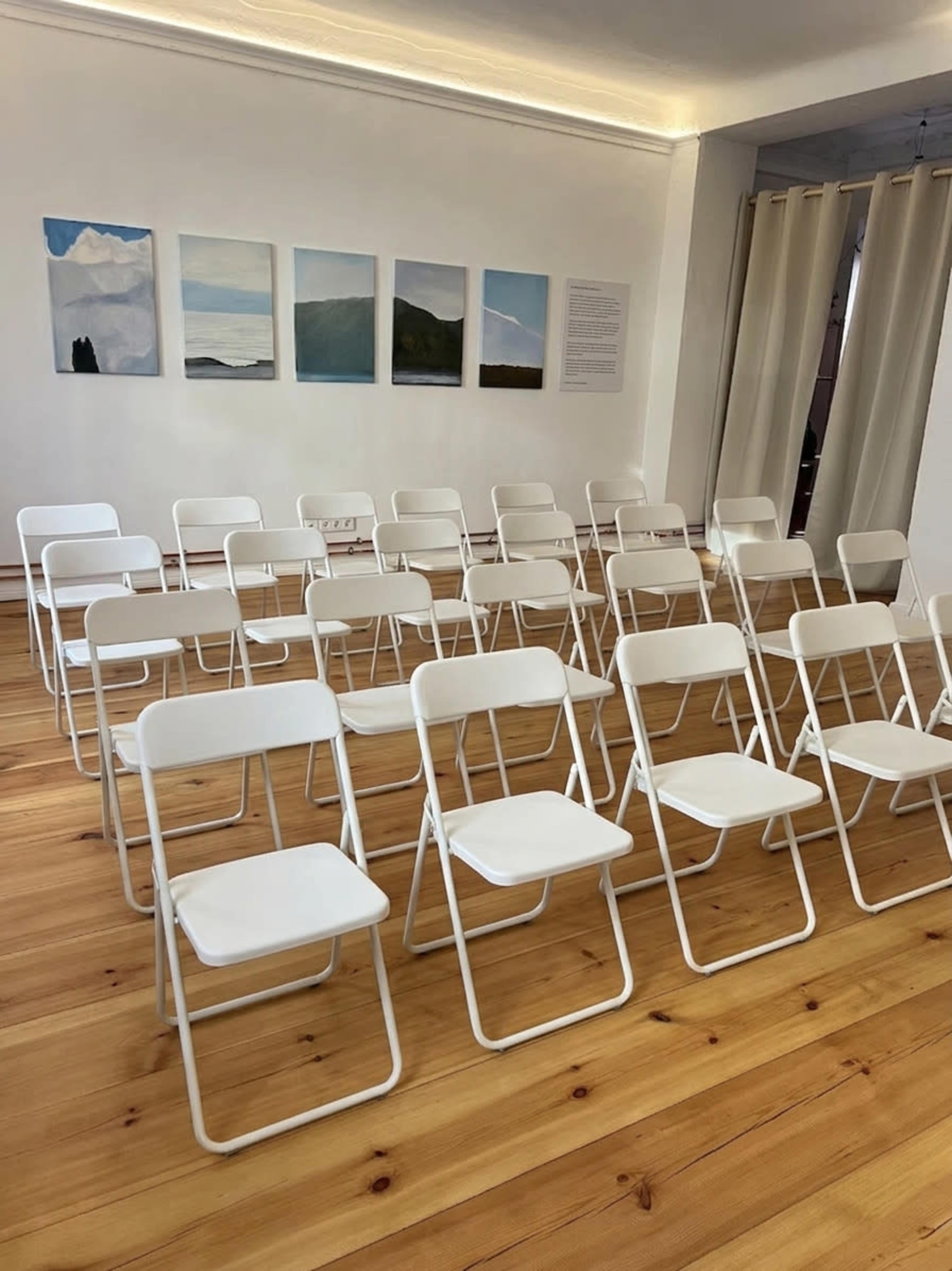 A row of white folding chairs is set up in a bright room with wooden flooring and artworks displayed on the walls.