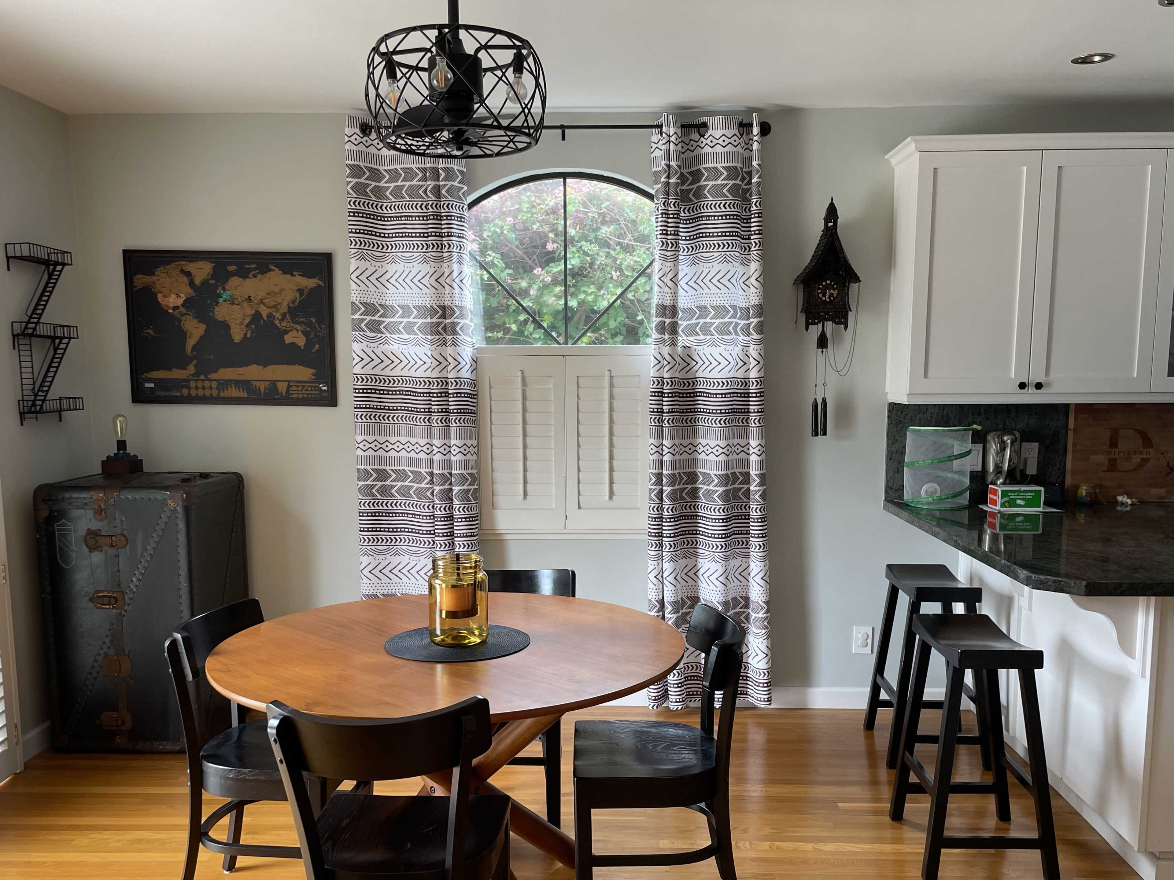 A round wooden table sits in a dining area with black chairs, adorned by patterned curtains and a map on the wall.