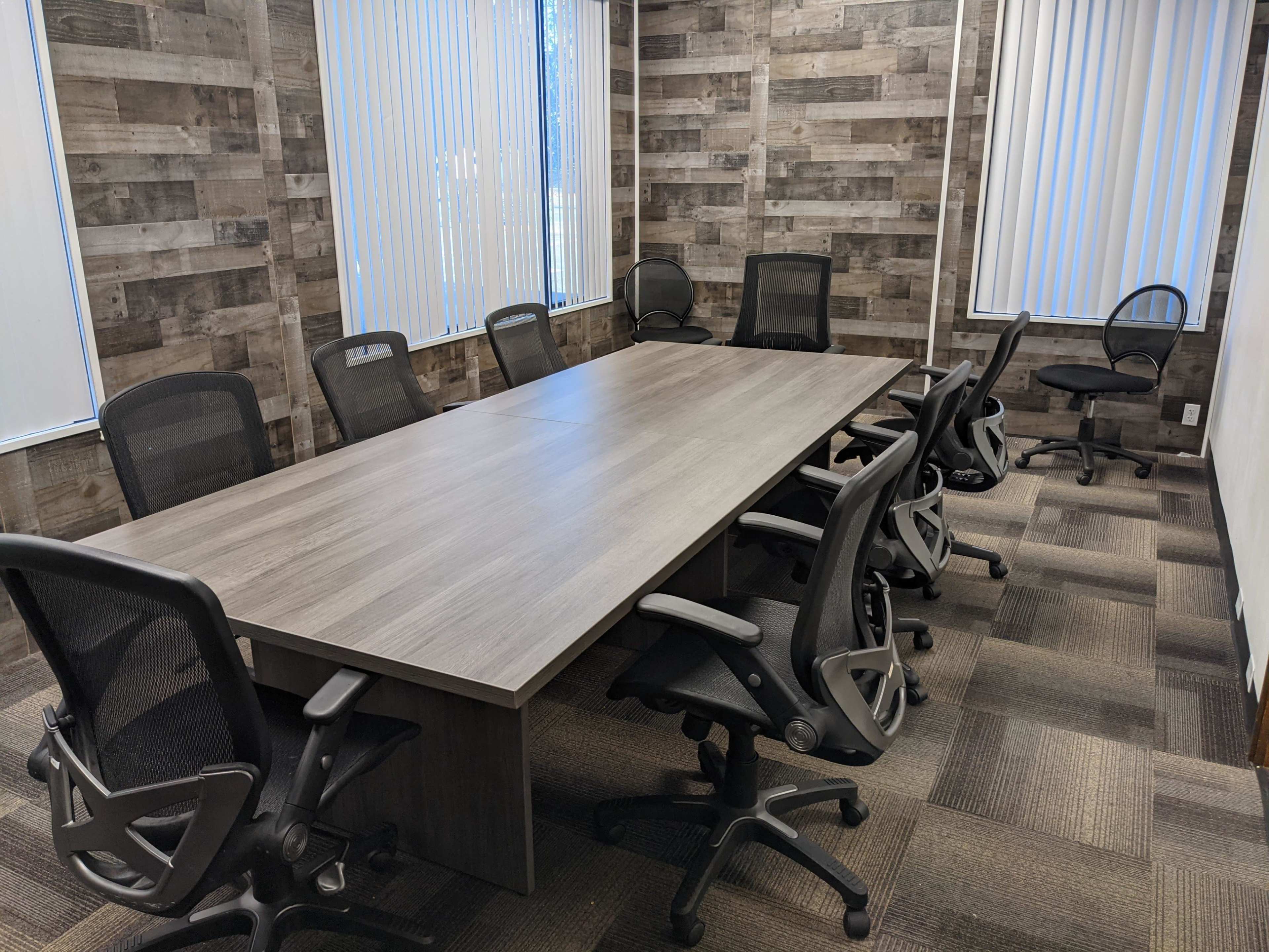 A large rectangular conference table surrounded by black office chairs is located in a room with wooden wall panels and vertical blinds.