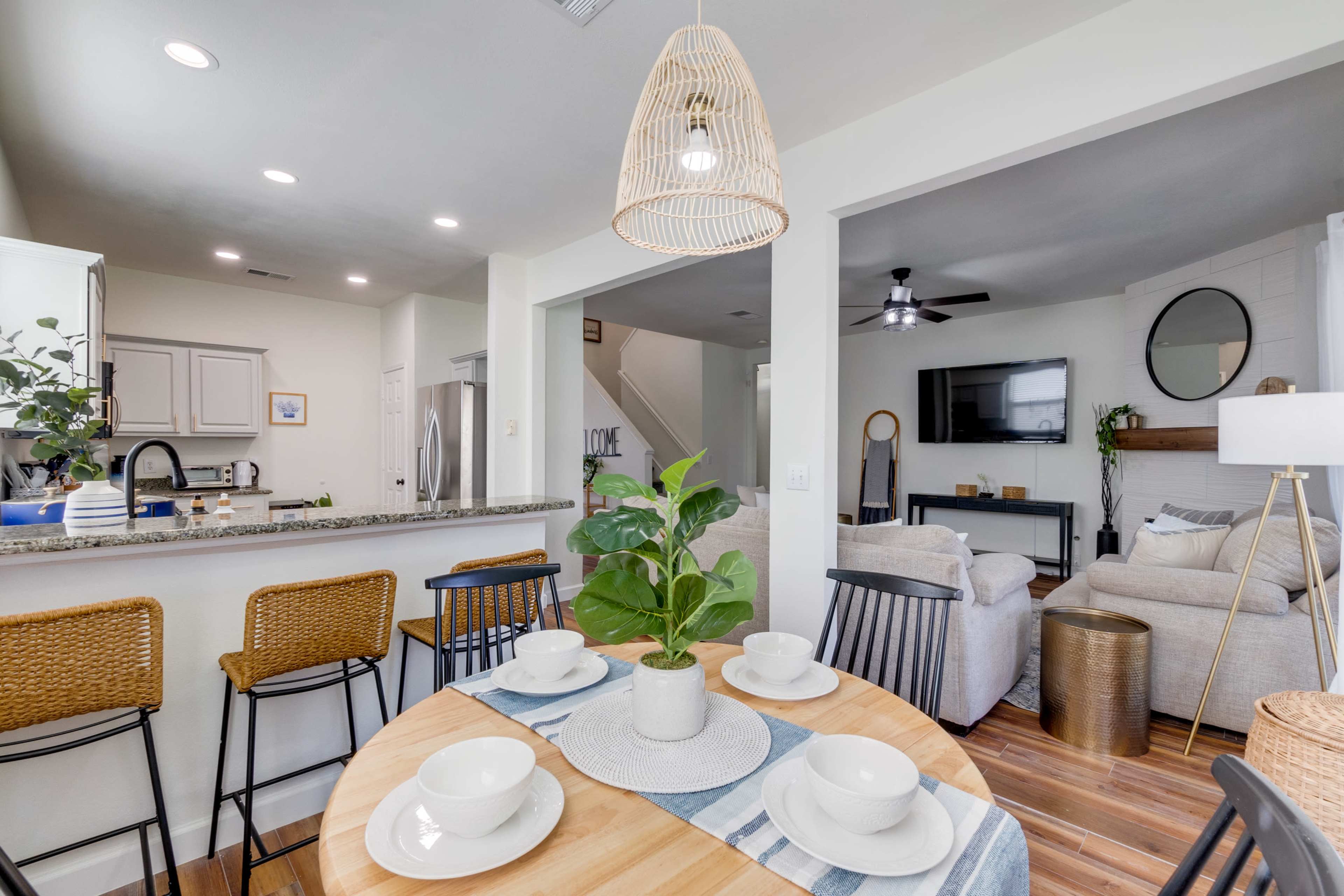 A dining area features a round wooden table set with dishware and a plant centerpiece, adjacent to a kitchen and living space with modern furnishings.