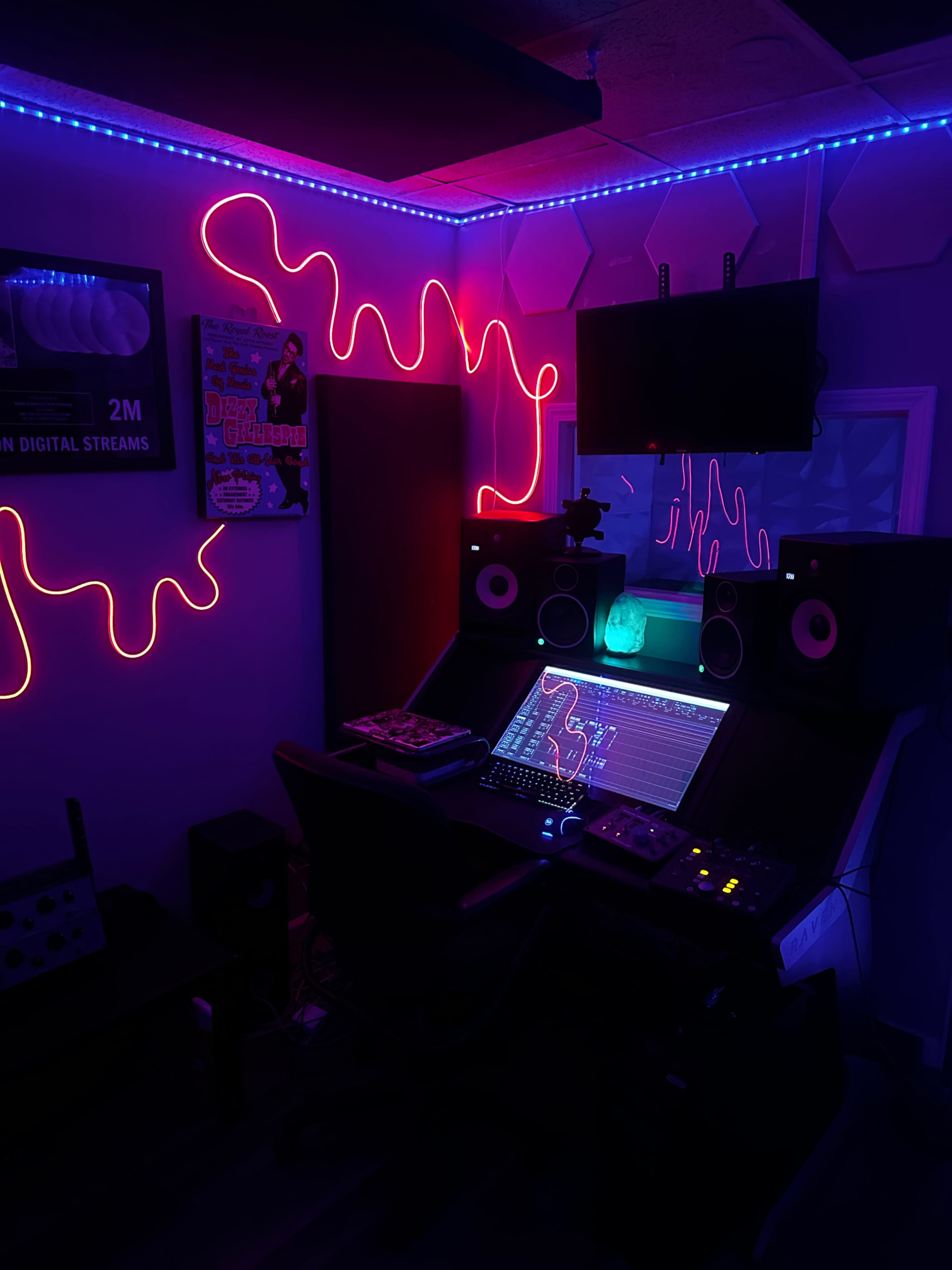 The image shows a dimly lit music studio featuring neon lighting, monitors, and audio equipment arranged on a central desk.