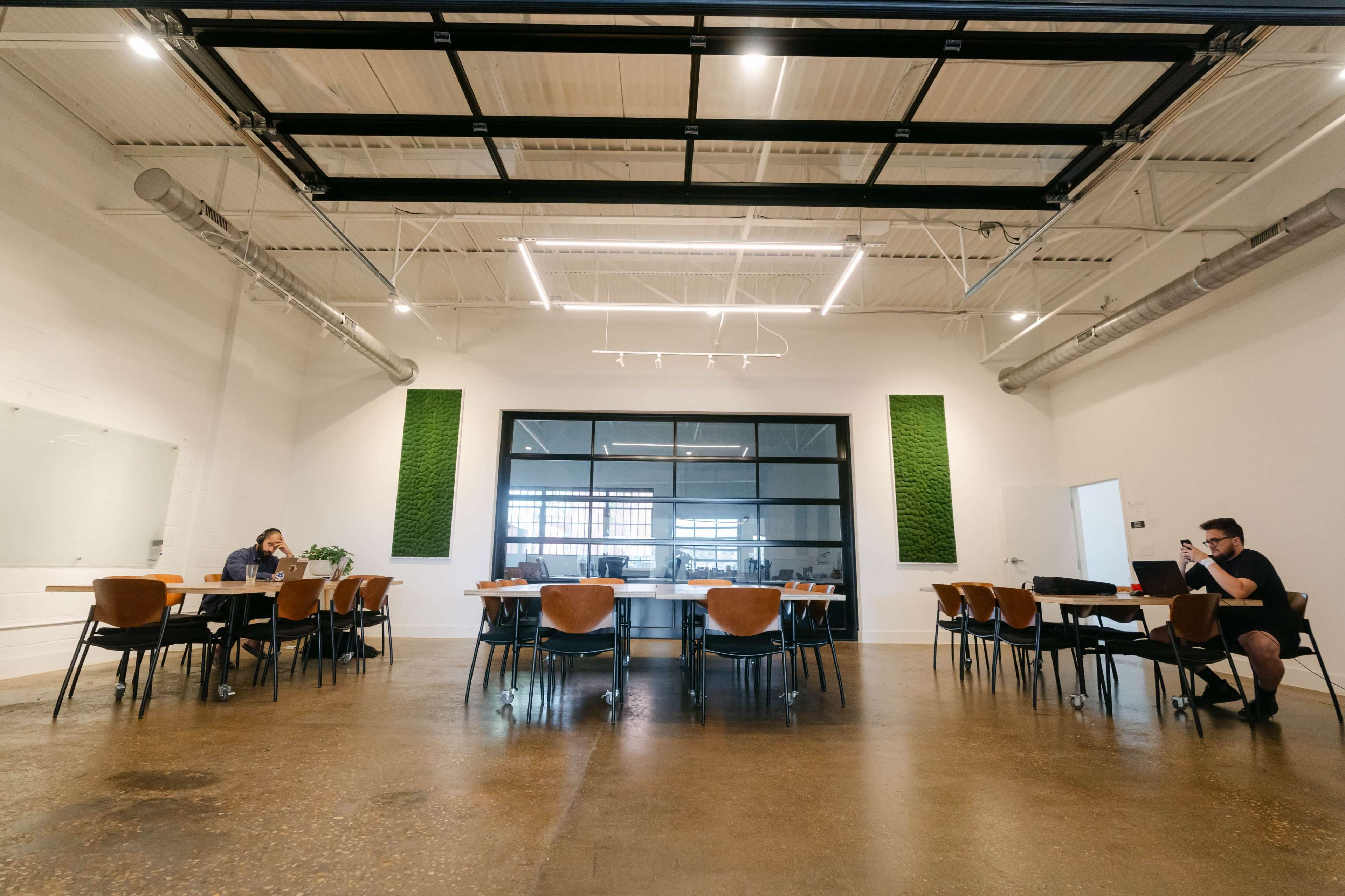 A spacious meeting room features a long table surrounded by chairs, with large windows and green wall accents.