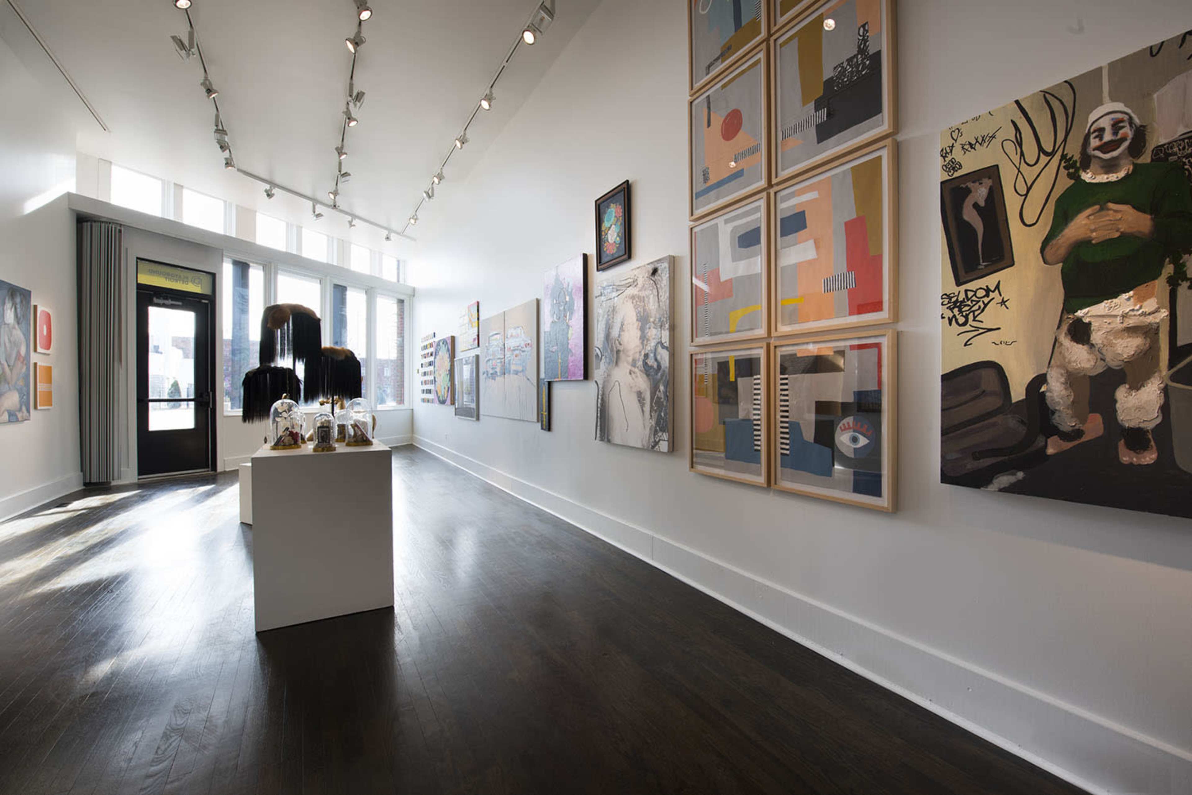 The image shows an art gallery with various colorful paintings displayed on the walls and a white pedestal holding art pieces in the center.