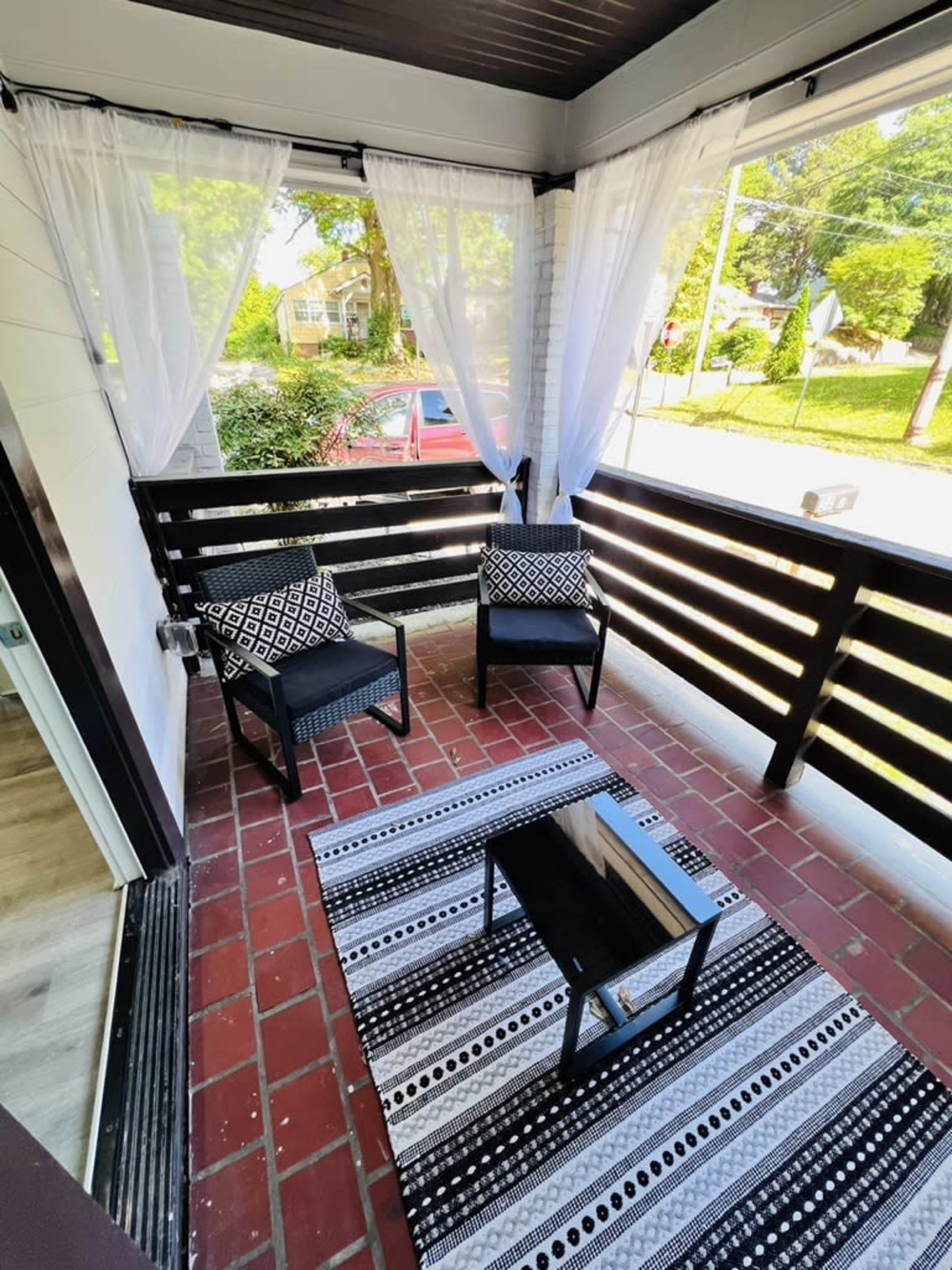 A cozy porch with two black chairs, a small table, and a patterned rug, framed by white sheer curtains.