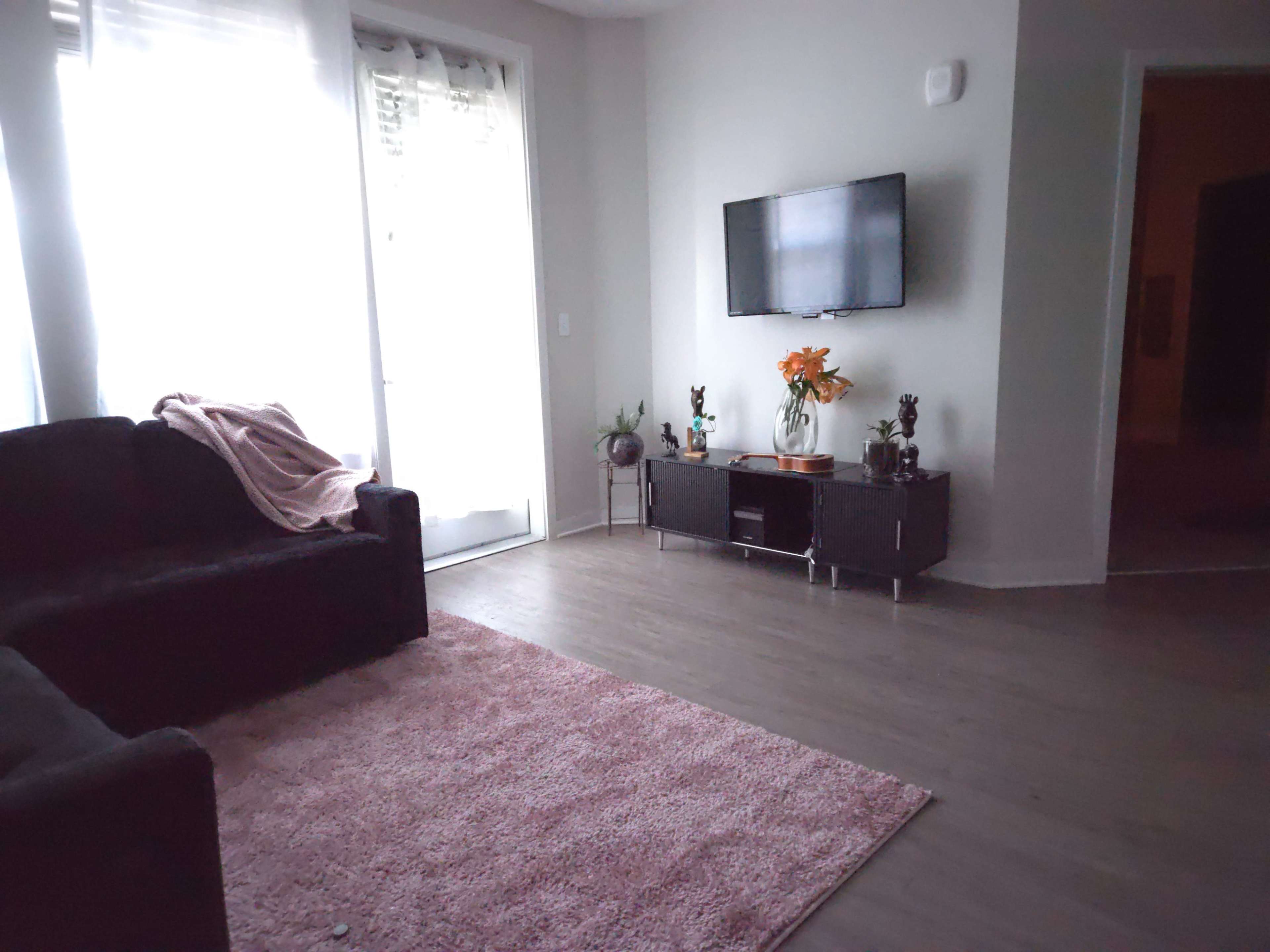 The image shows a modern living room featuring a black couch, a television mounted on the wall, and a console with decorative items and flowers beside a large window.