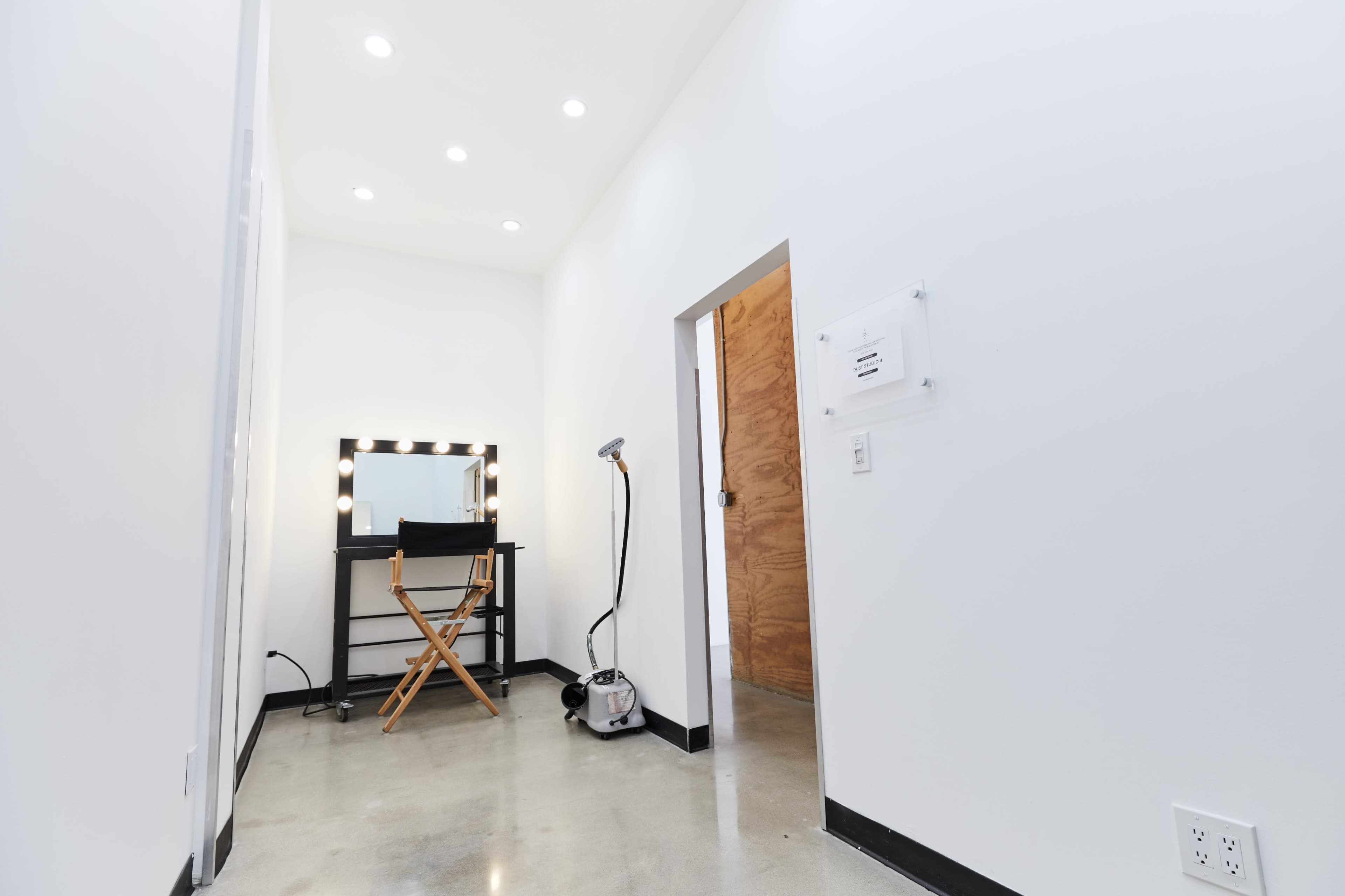 The image shows a brightly lit, minimalist hallway leading to a room with a makeup vanity and a director's chair.