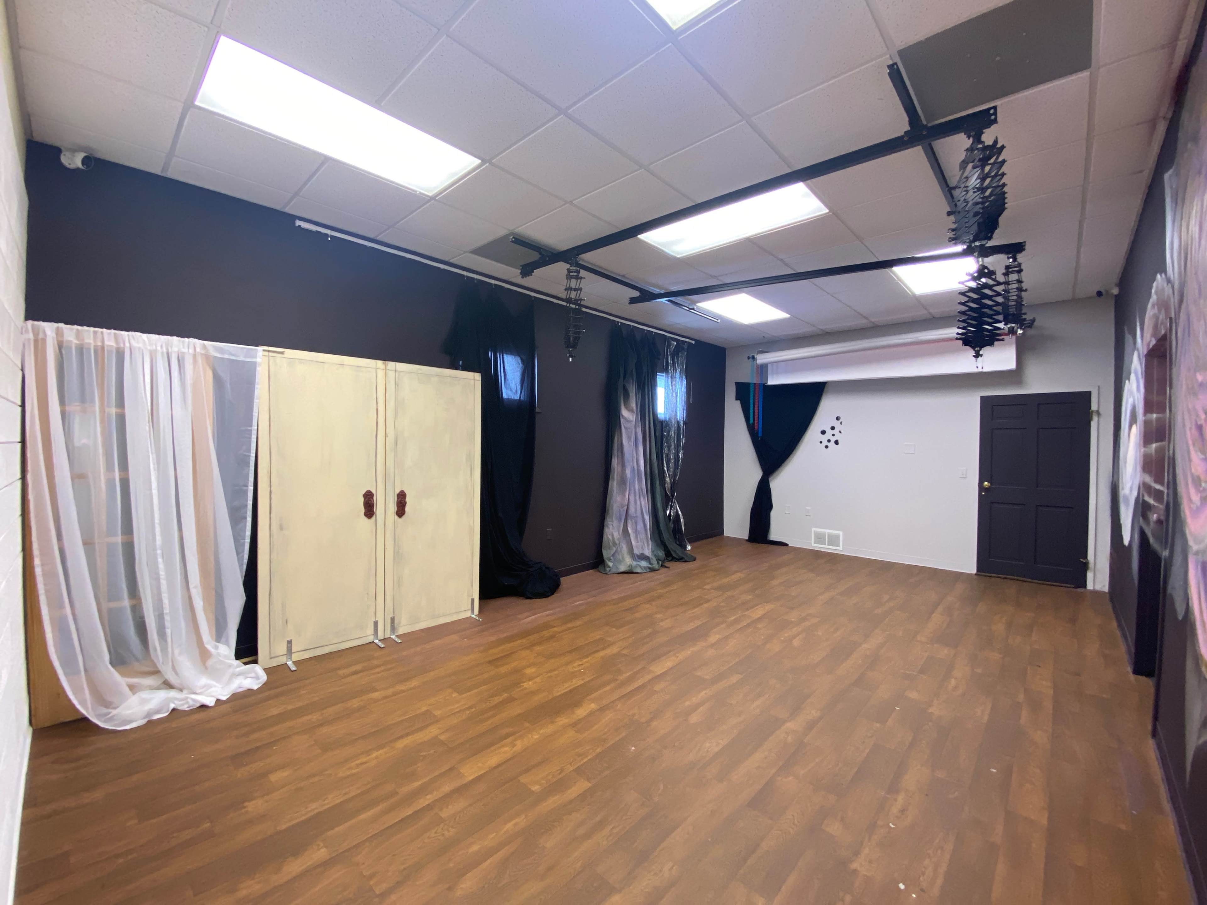 A spacious room with wooden flooring, featuring a light-colored wardrobe, black curtains, and several windows.