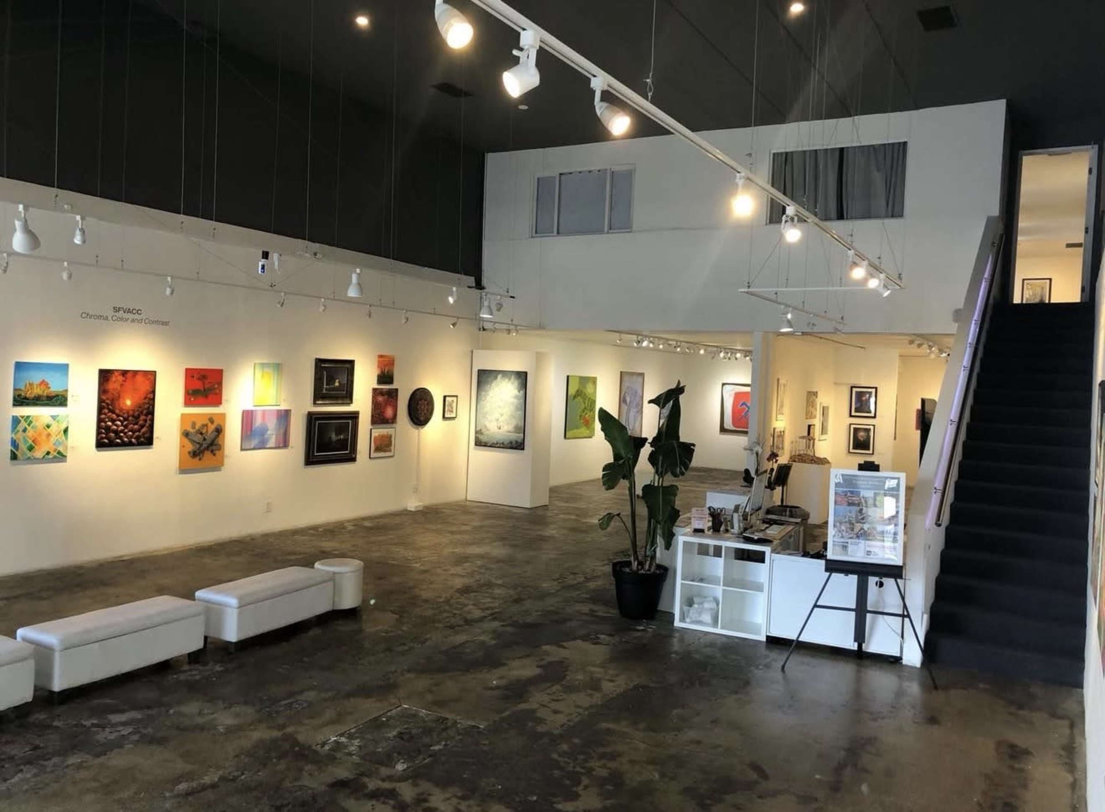 The image shows an art gallery with various paintings displayed on the white walls, a reception area with a desk, and stairs leading to an upper level.