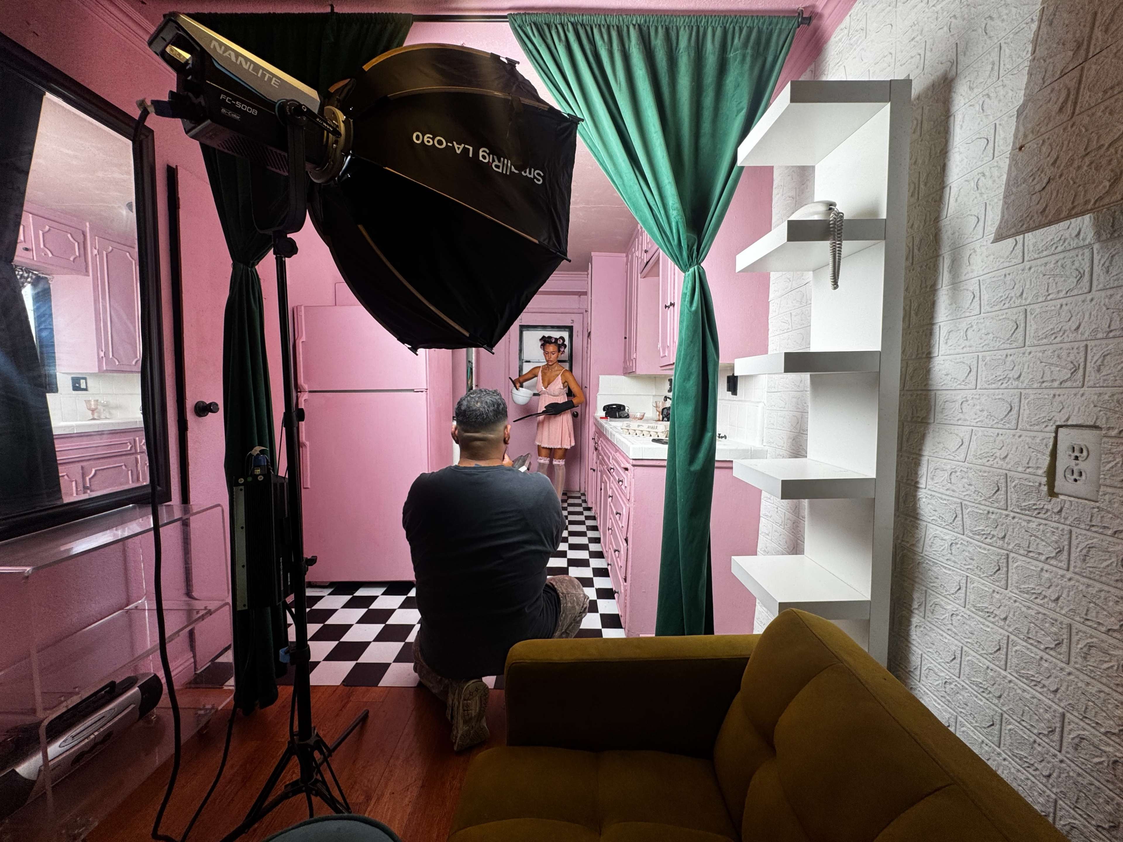 A photographer captures a model in a pink and black checkered kitchen setting, with lighting equipment positioned nearby.