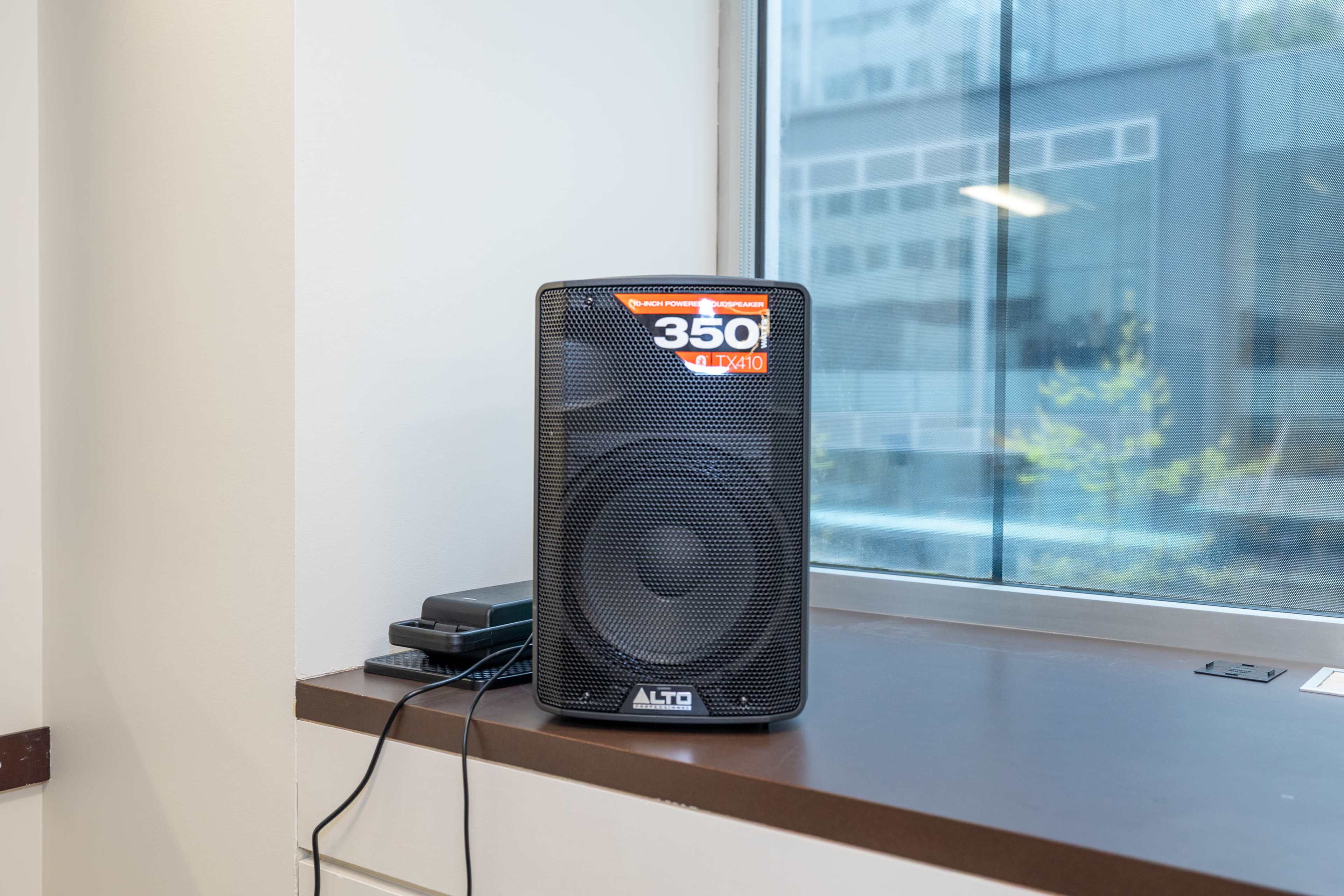 A black Alto speaker sits on a windowsill next to a small electronic device, with a cityscape visible through the window.
