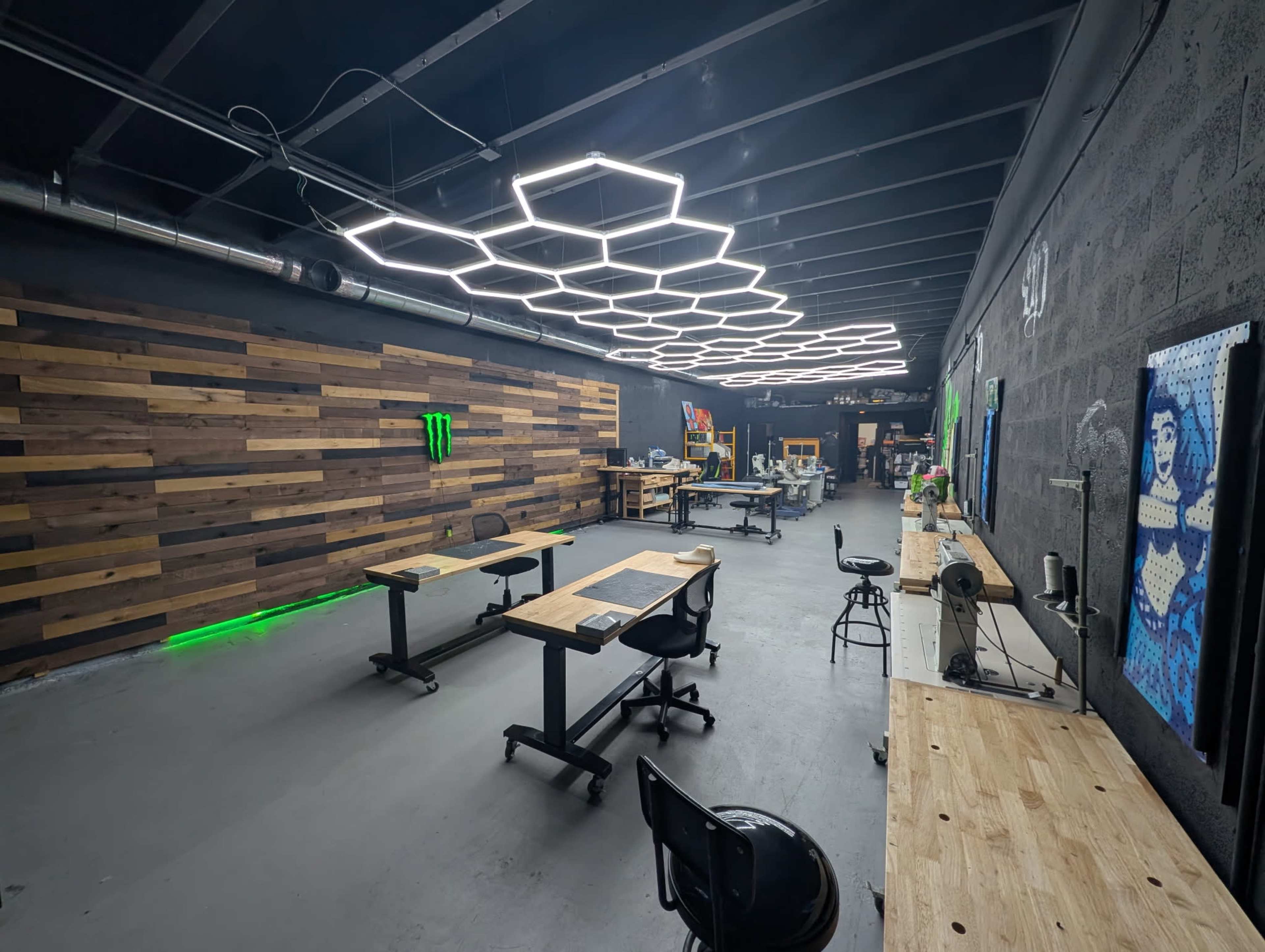 The image shows a spacious workshop with wooden tables, industrial lighting in a hexagonal pattern, and a wooden accent wall, featuring a green Monster logo.