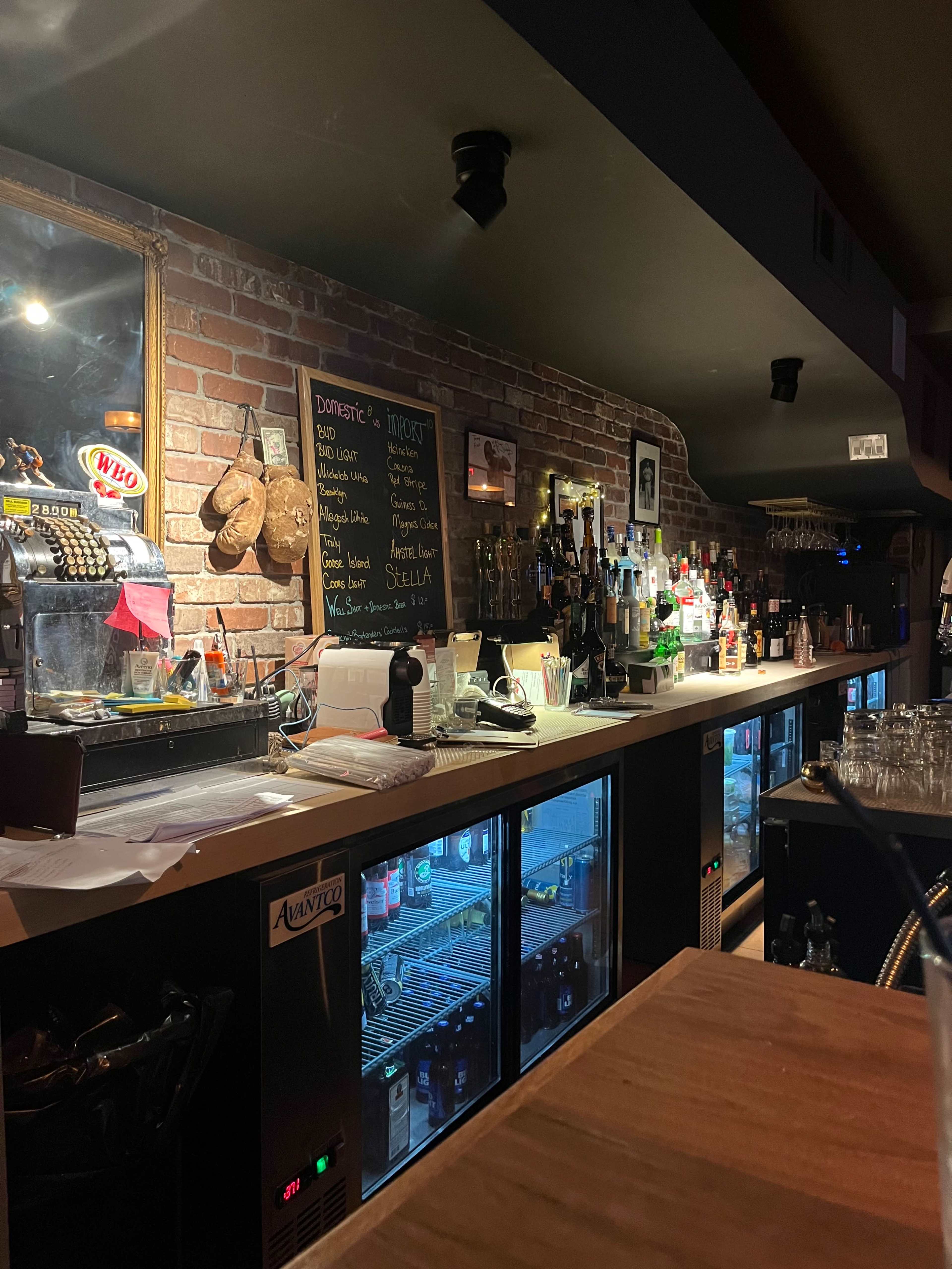 A dimly lit bar with a wooden counter, exposed brick walls, and a variety of liquor bottles displayed on shelves behind the bar.